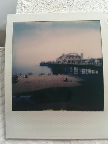 Polaroid photo: The Brighton Palace pier jets out to sea . A white structure on metal pillars and frame visible underneath. On top is the dome where the arcade machines are. In the foreground are people enjoying the pebble beach and the sunny day.