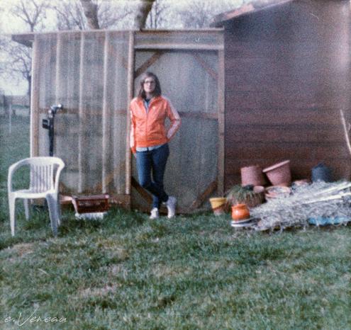 A woman in an orange jacket stands leaning against the door of a garden shed. You can make out the trees behind. In front: a white plastic chair and empty flower pots.