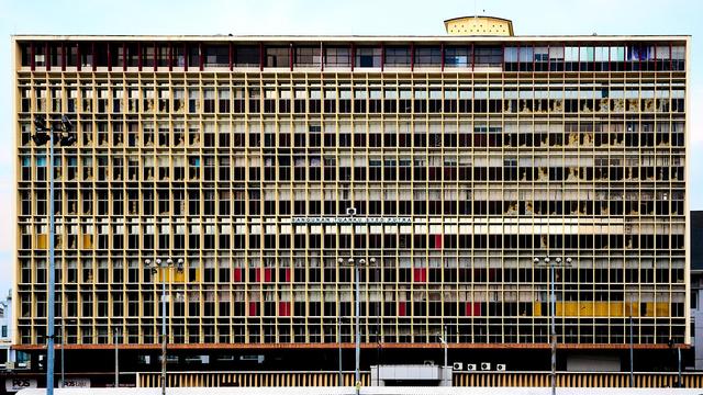 The image shows a large rectangular building with a grid-like facade of numerous windows. The structure has a flat roof and a sign with the name 'Bangunan Tuanku Syed Putra.' Several lamp posts are visible in front of the building