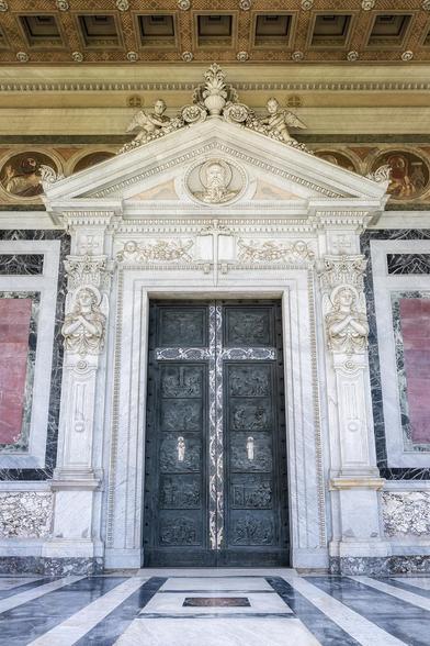 The ornate, intricately carved main doorway of the Basilica of St. Paul Outside The Walls is made of bronze with silver inlays. It is flanked by marble sculptures and framed by an intricately detailed pediment with classical motifs. 