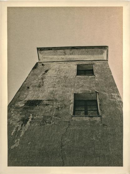 This image is a lith print made on old photographic paper. It depicts an upward perspective of a weathered, tall concrete or stucco building with visible surface cracks and peeling paint. The composition emphasizes the verticality of the structure, with two dark, rectangular windows interrupting the facade. The sky above is featureless and pale, adding to the stark and minimalist atmosphere. The lith printing process gives the photograph a distinctive tonal quality—rich, gritty blacks with a warm, slightly sepia-toned midtone and highlight range, contributing to its vintage, textured aesthetic. The paper’s aged look and the unique grain from the lith technique enhance the overall expressive mood of the print.
