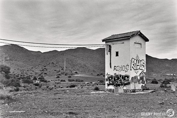 An electrical sub-station with graffiti stands in a barren, rocky landscape. Mountains loom in the background under a cloudy sky. Captured with a Nikon F on Ilford Delta 100 film, power lines spur across the scene.