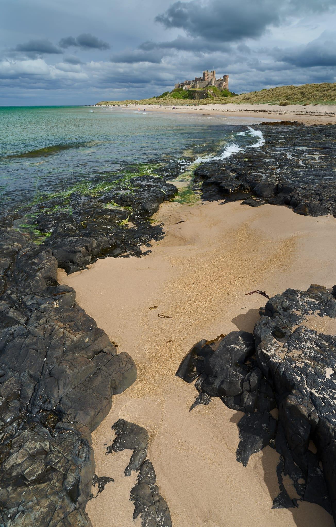 A dramatic view of Bamburgh Castle sits atop grassy dunes in the distance, overlooking a sweeping sandy beach edged by jagged dark rocks and patches of green seaweed, with turquoise waves gently lapping the shore beneath a moody, cloud-filled sky.
