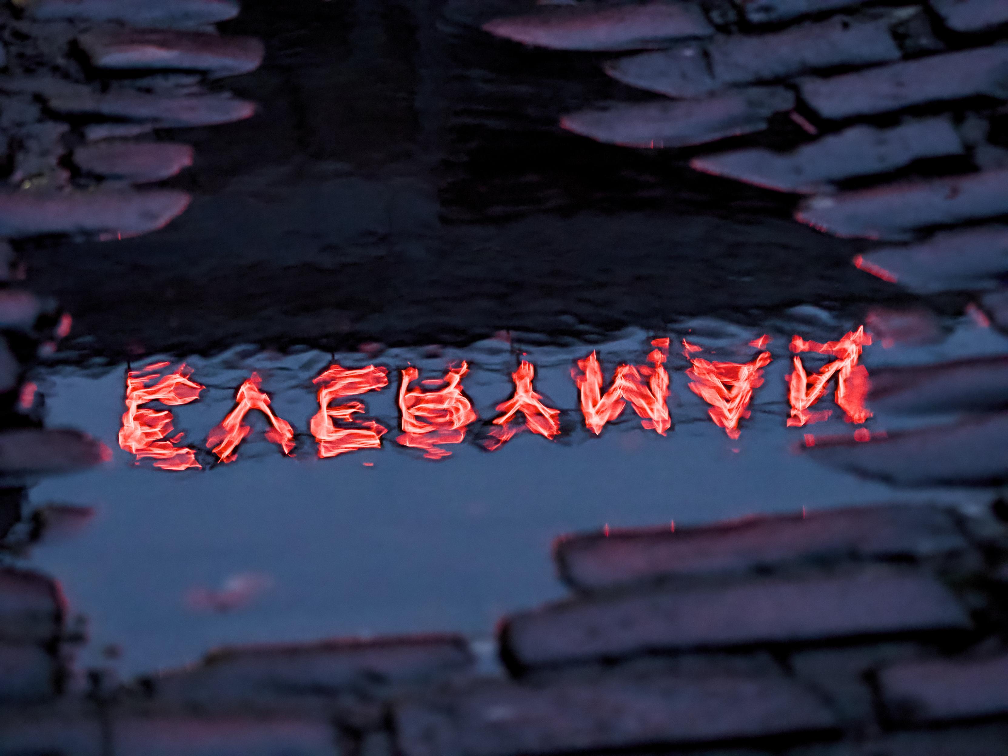 The word EVERYMAN, written in red neon capitals, reflects in a puddle among cobbled stones in an alleyway, the letters are reversed and shimmering because of a light breeze rippling the surface of the water