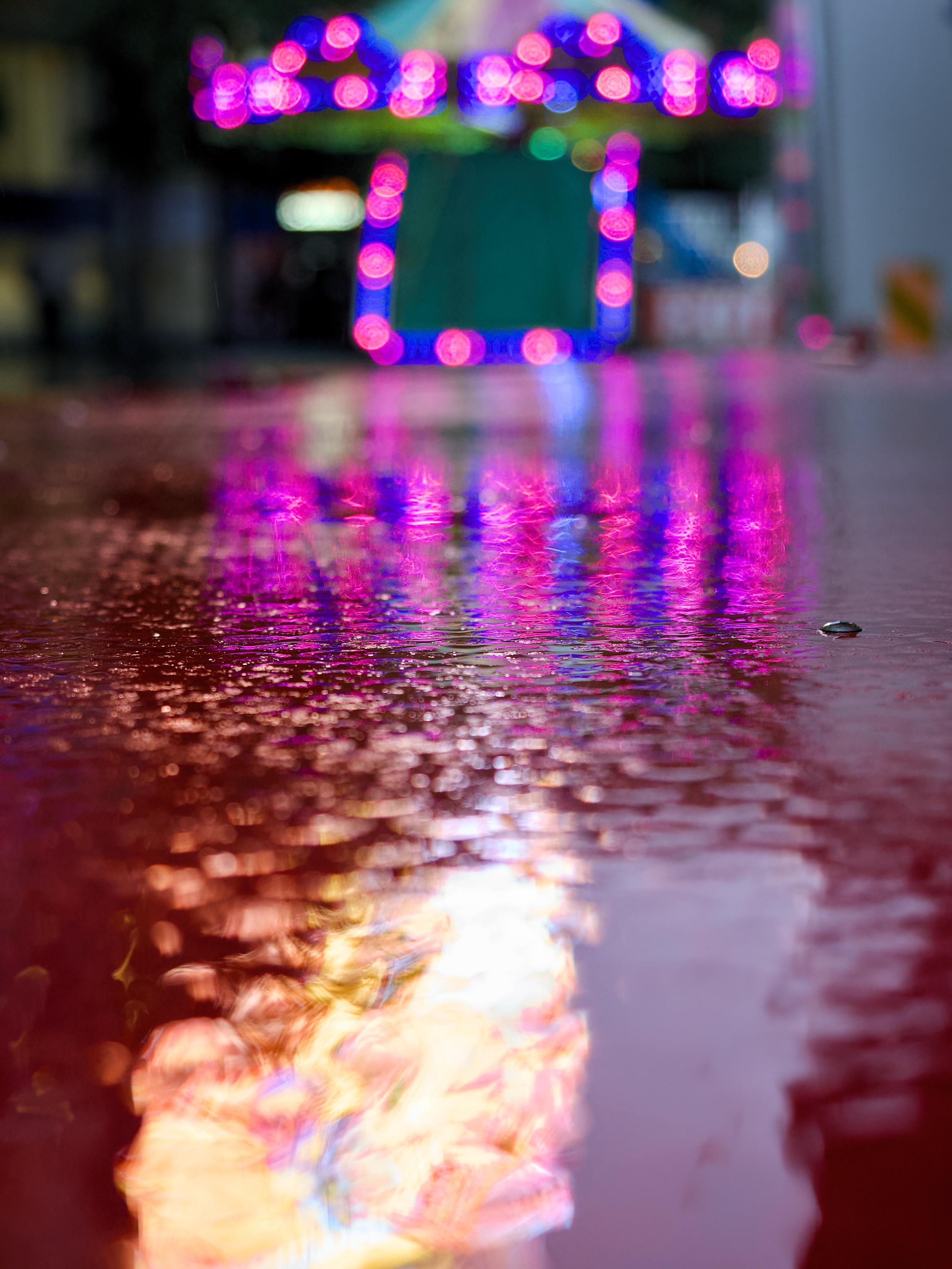 The metallic surface of a fairground ride reflects pink and blue neon lights in the background, along with the flame-like reflections from a neon advertising board in the foreground.