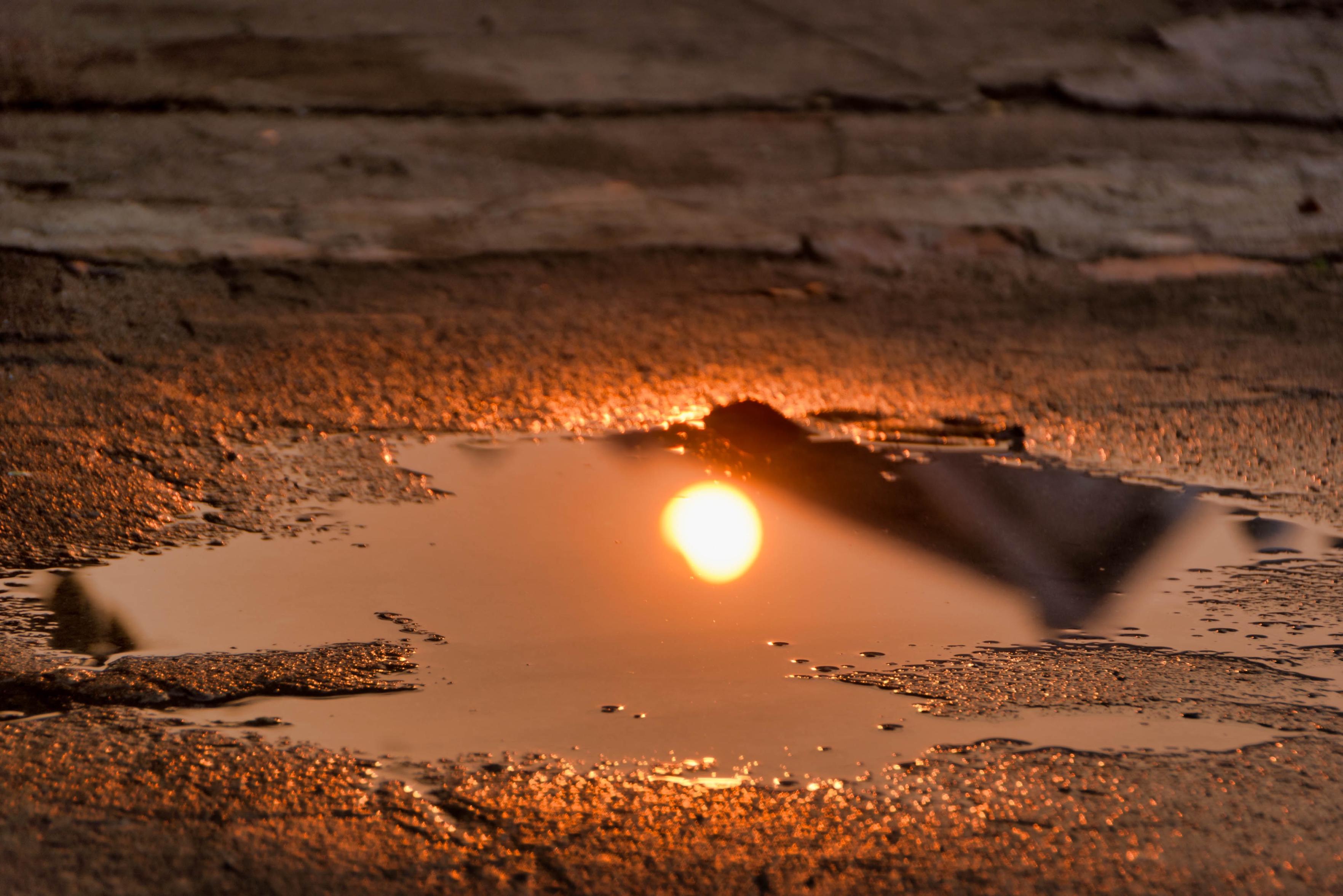 A shallow puddle reflecting the sunset, with shimmering orange hues on the water's surface and textured ground surrounding it. A shallow puddle reflecting the sunset, with shimmering orange hues on the water's surface and textured ground surrounding it.