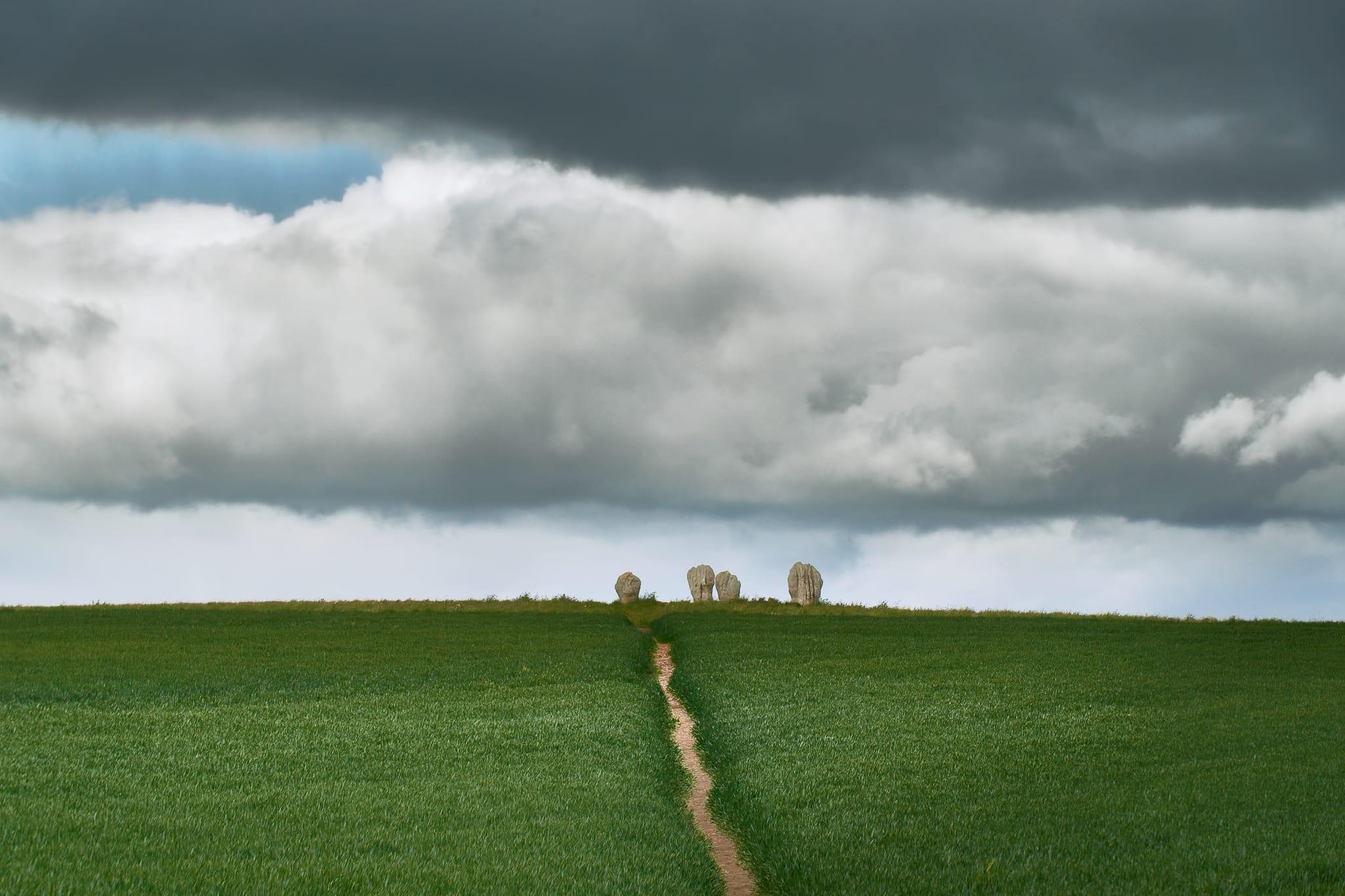 The Duddo Five Stones, a small group of ancient standing stones set on a gentle grassy hill in Northumberland, England. A narrow dirt path leads straight through the lush green field, drawing the eye towards the stones standing silhouetted against a dramatic sky filled with thick, low-hanging grey clouds. The stones appear weathered and irregular in shape, emphasising their age and historical significance, while the moody sky and open landscape evoke a sense of timelessness and mystery.