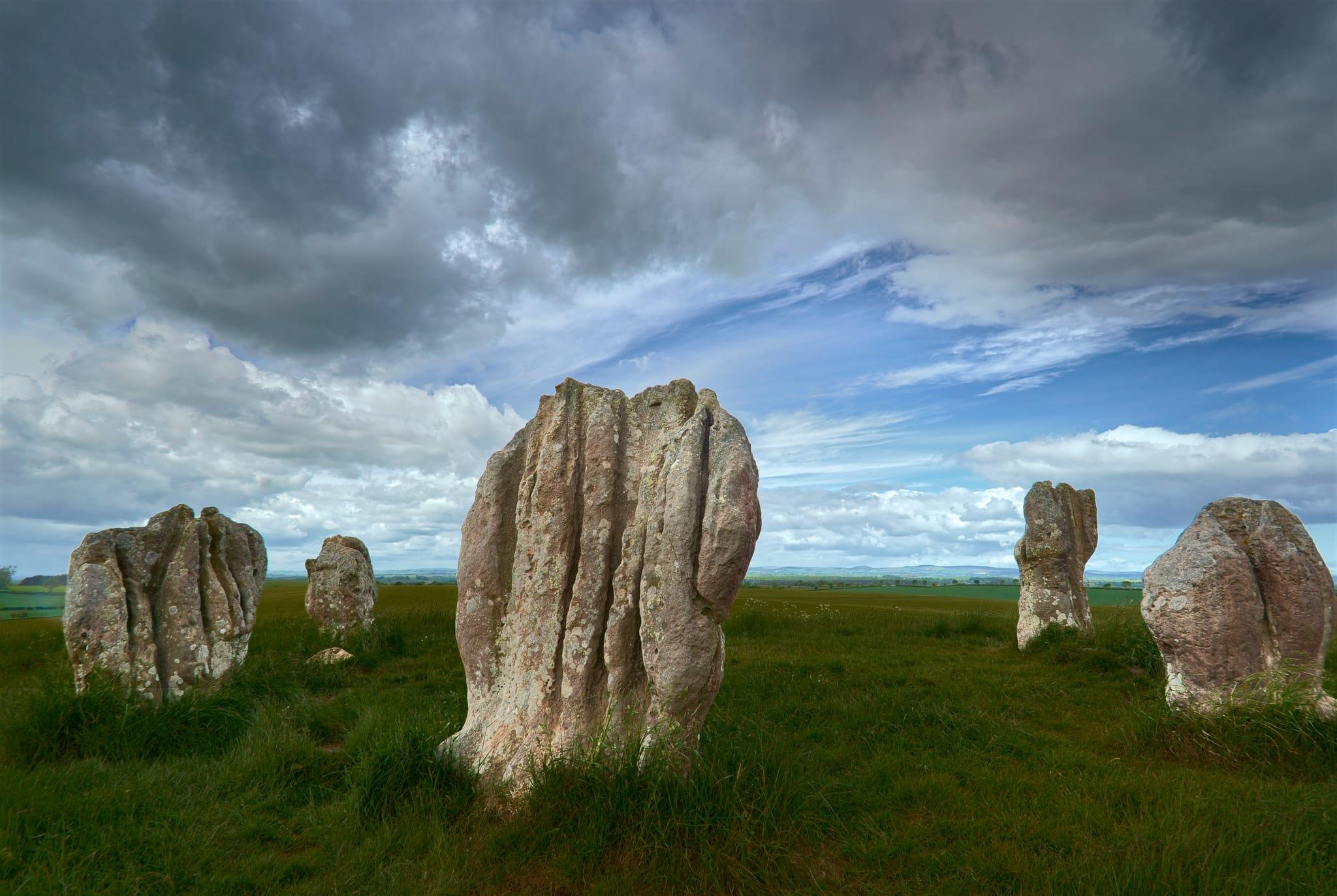 A close-up view of the Duddo Five Stones in Northumberland, England, showcasing the five large, weathered standing stones arranged in a rough circle on a grassy hilltop. The stones are irregular in shape, covered with patches of lichen, and their rough textures and vertical grooves are clearly visible. The surrounding grass is lush and green, while the sky above is dynamic with a mix of dark, heavy clouds and patches of blue sky, casting a moody yet vibrant atmosphere over the ancient site and its expansive rural landscape in the distance.