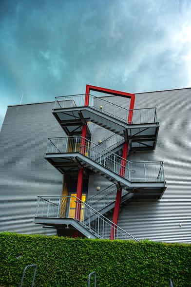 Gebäude mit aus Wellblechplatten verkleideter Außenfassade. An diese ist eine Treppenanlage über drei Stockwerke angesetzt. Im Vordergrund steht eine akkurat geschnittene Hecke.