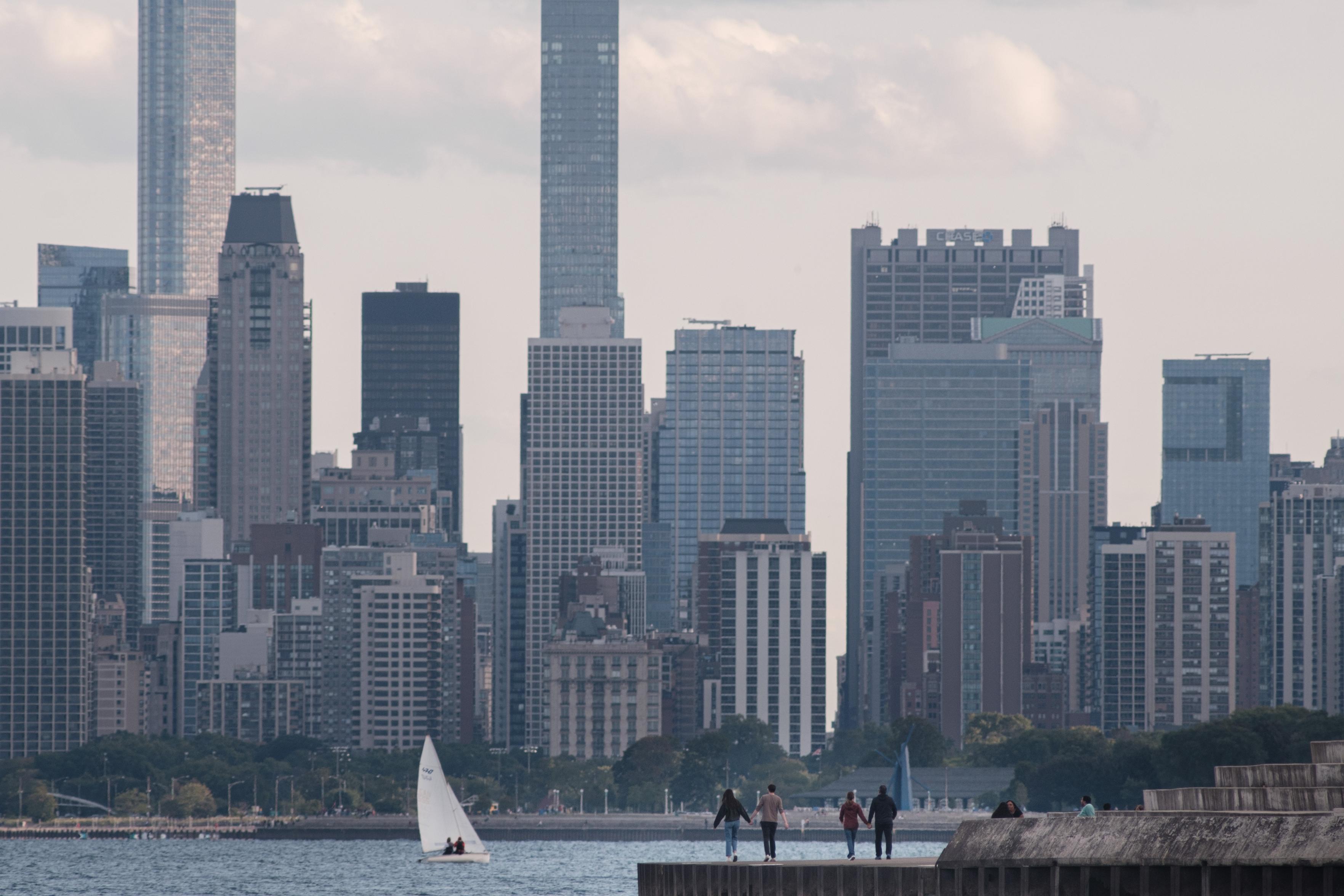 Two couples walk along a concrete path next to Lake Michigan while holding hands as a sailboat passes behind them. The building of downtown Chicago are present in the background.