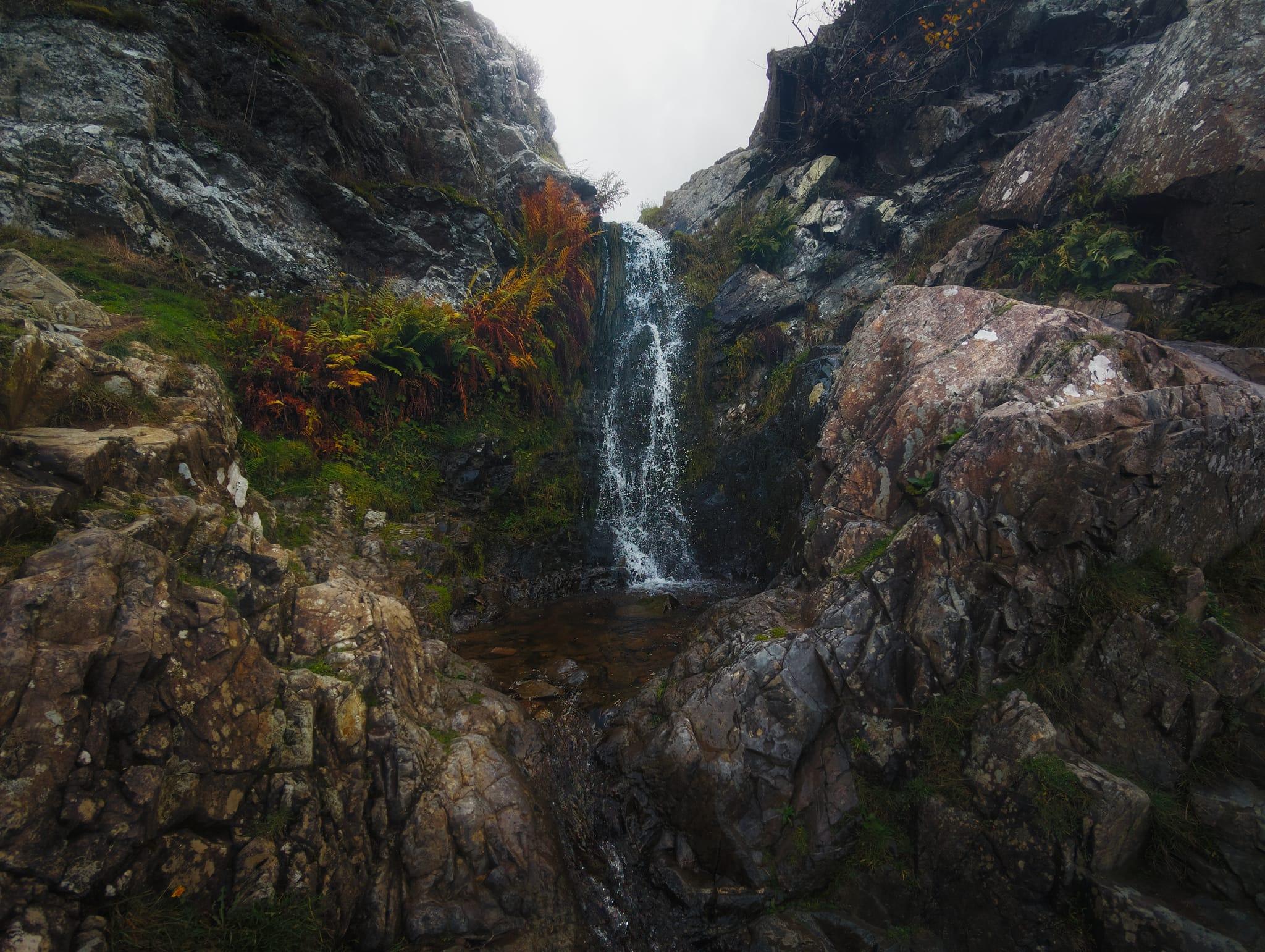 A narrow waterfall cascades down a dark rocky cliff face into a small dark pool within a dramatic gorge, flanked by steep stratified limestone walls covered in moss, lichen, and autumn vegetation in shades of burnt orange, rust, and sage green. The waterfall's white water contrasts sharply against the shadowed stone as it tumbles between the towering rock faces, which display natural geological layering and weathered surfaces stained with mineral deposits and algae growth. Boulders and rocky outcrops jut from the ground in the foreground, their surfaces worn smooth by water and time, whilst overhanging vegetation and ferns cling to crevices along the cliff edges, creating an intimate and secluded natural amphitheatre typical of the hidden geological gems found throughout the Shropshire Hills.