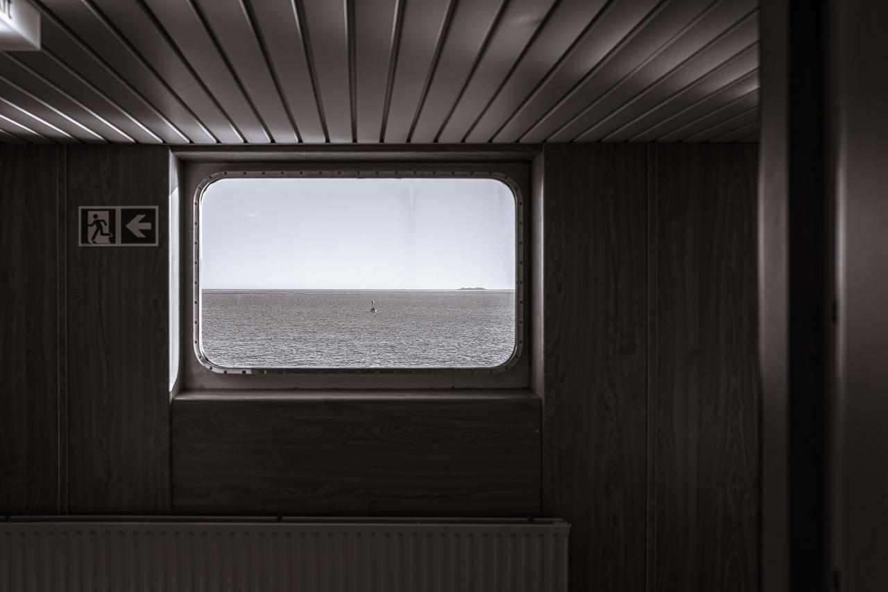Window in a ferry en route to North Sea Island Amurm - through the window a buoyage and a small island in the calm sea is visible. Above the window there are reflections of sunlight illuminating the ceiling, below the window.a part of a radiator is visible