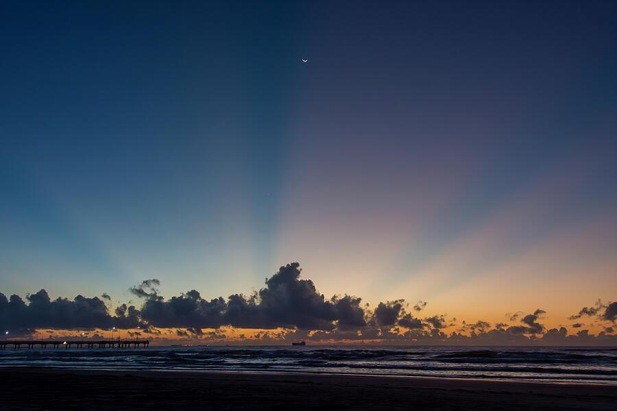 The sky is painted in hues of orange and blue as the sun rises above the ocean. Clouds and a moon crescent create a stunning silhouette against the light. ©Debra Martz, All Rights Reserved