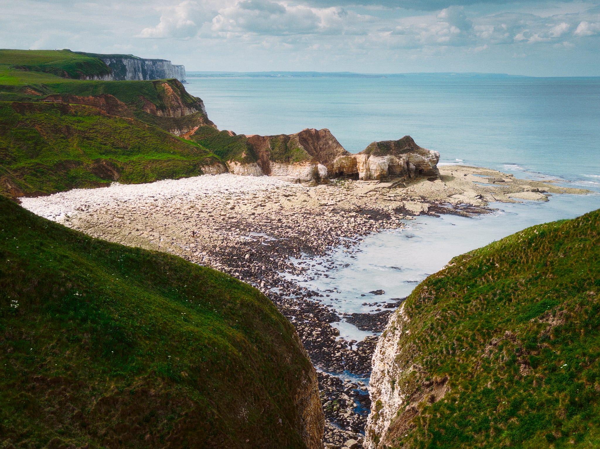 View from the cliffs overlooking Thornwick Bay on the Flamborough coastline in East Riding of Yorkshire, with steep green headlands surrounding a broad rocky beach and pale chalk formations jutting into the calm blue sea beneath a partly cloudy sky.