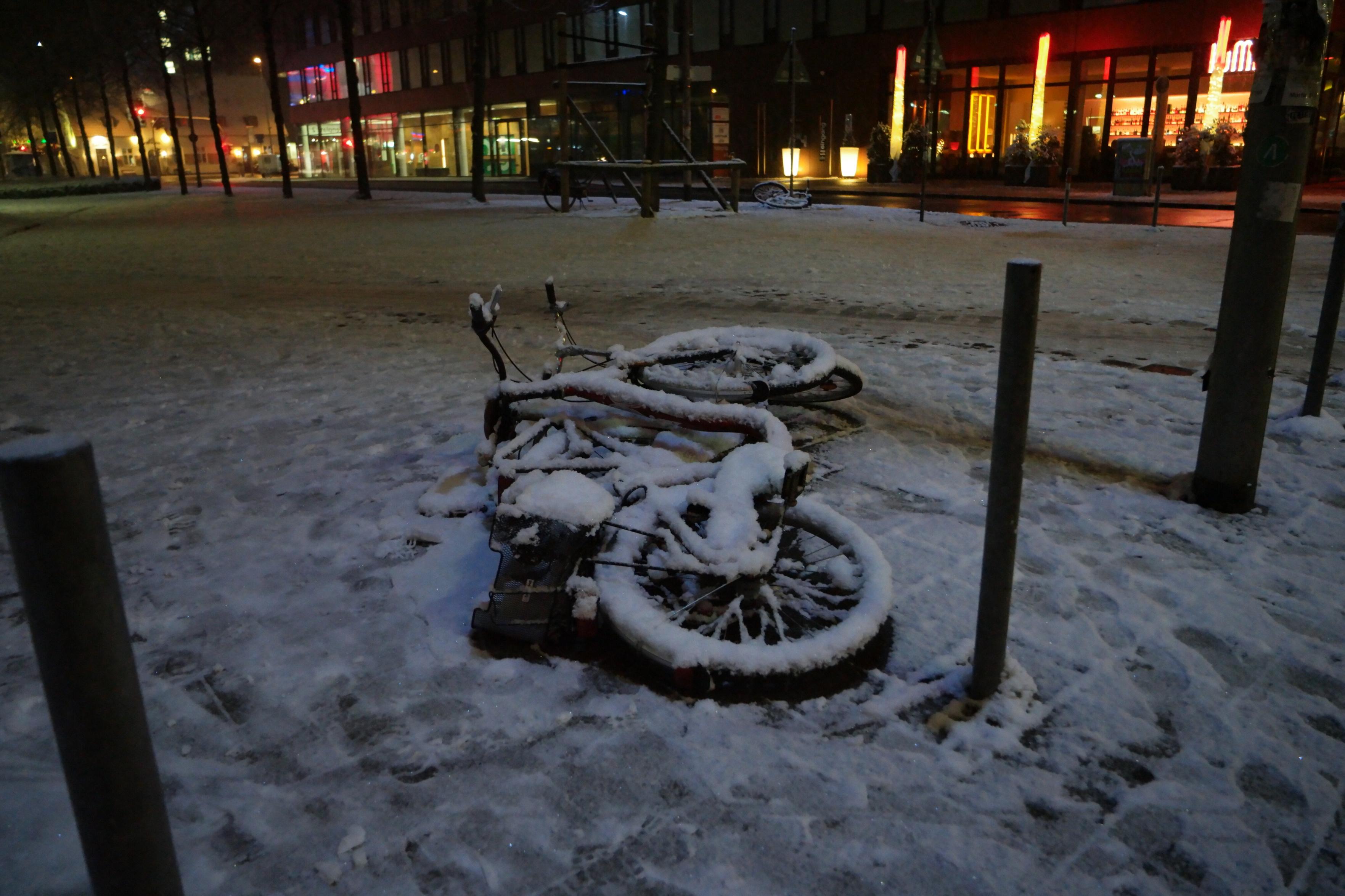 Winterfoto: zwei Fahrräder liegen umgekippt neben einem Pfahl auf einem weitläufigen Platz. Die Fahrräder und der Platz sind mit einer Schicht Schnee bedeckt. Im Hintergrund sind die Lichter einer Einkaufsmeile zu sehen