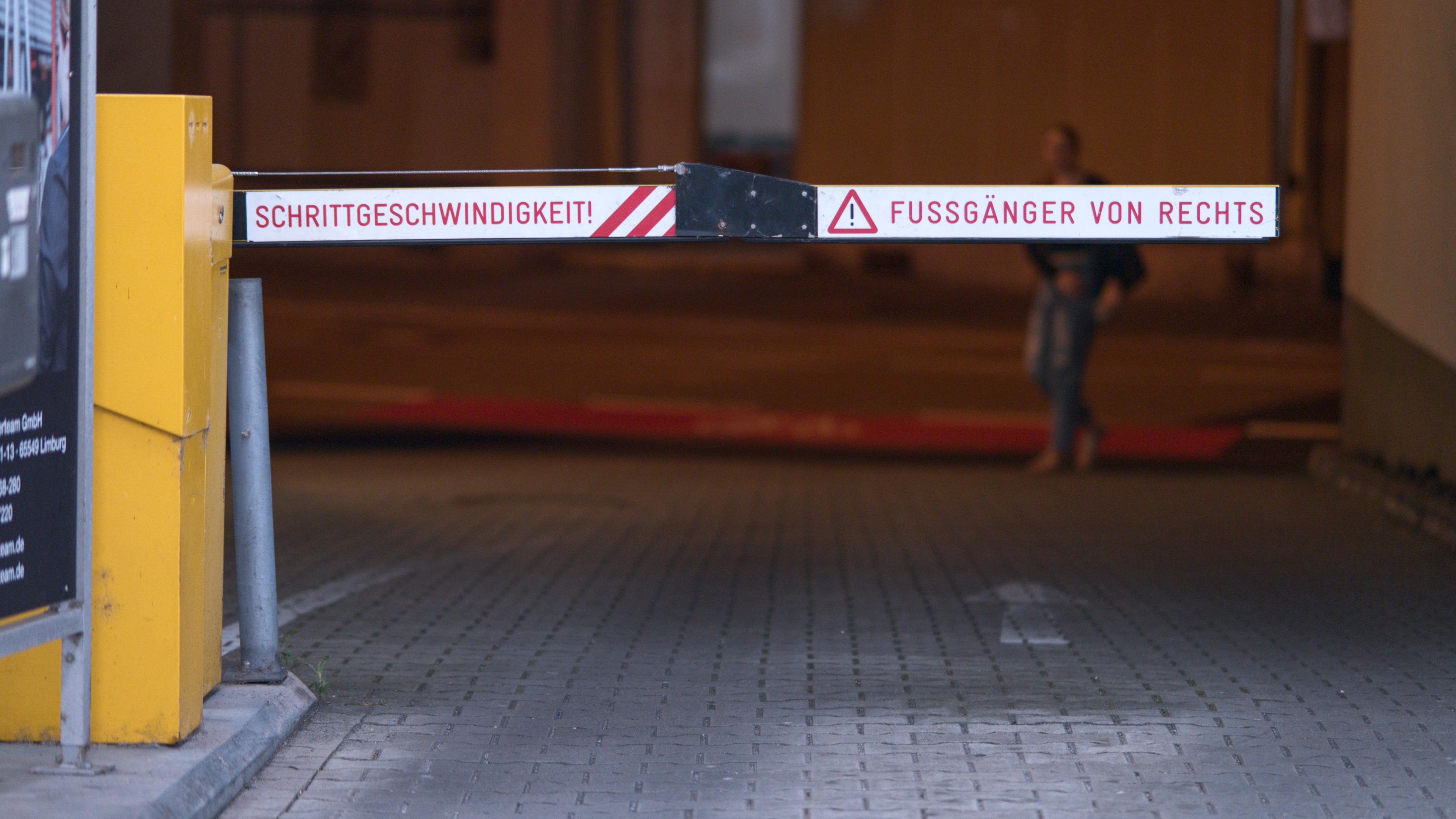 Ausfahrt aus einem Parkhaus von innen. Eine Schranke versperrt den Weg. Auf der Schranke ist in rot auf reflektierend weißem Grund geschrieben: "Schrittgeschwindigkeit! Fußgänger von rechts". Im Hintergrund hinter der Schranke läuft eine Person von rechts zur Bildmitte.