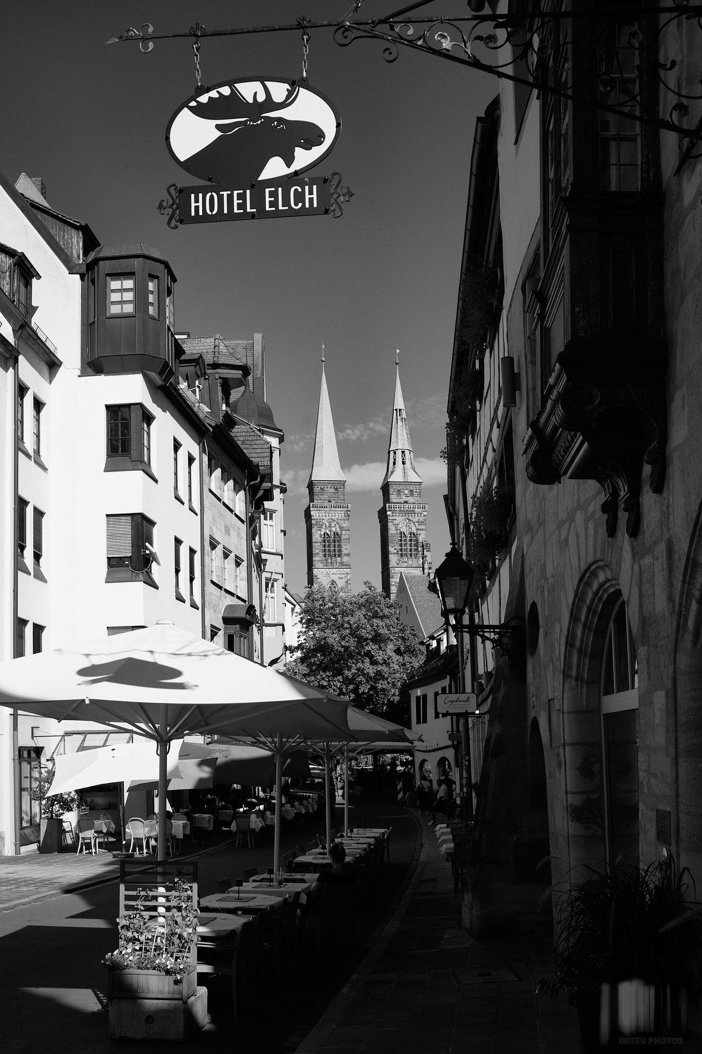 Black and white street photograph in Nuremberg's medieval old town. A wrought-iron 'Hotel Elch' sign with an elk emblem hangs over a narrow lane in the foreground, café tables and umbrellas below. The lane curves back toward the twin Gothic spires of St. Sebald's Church rising above half-timbered facades in the background.