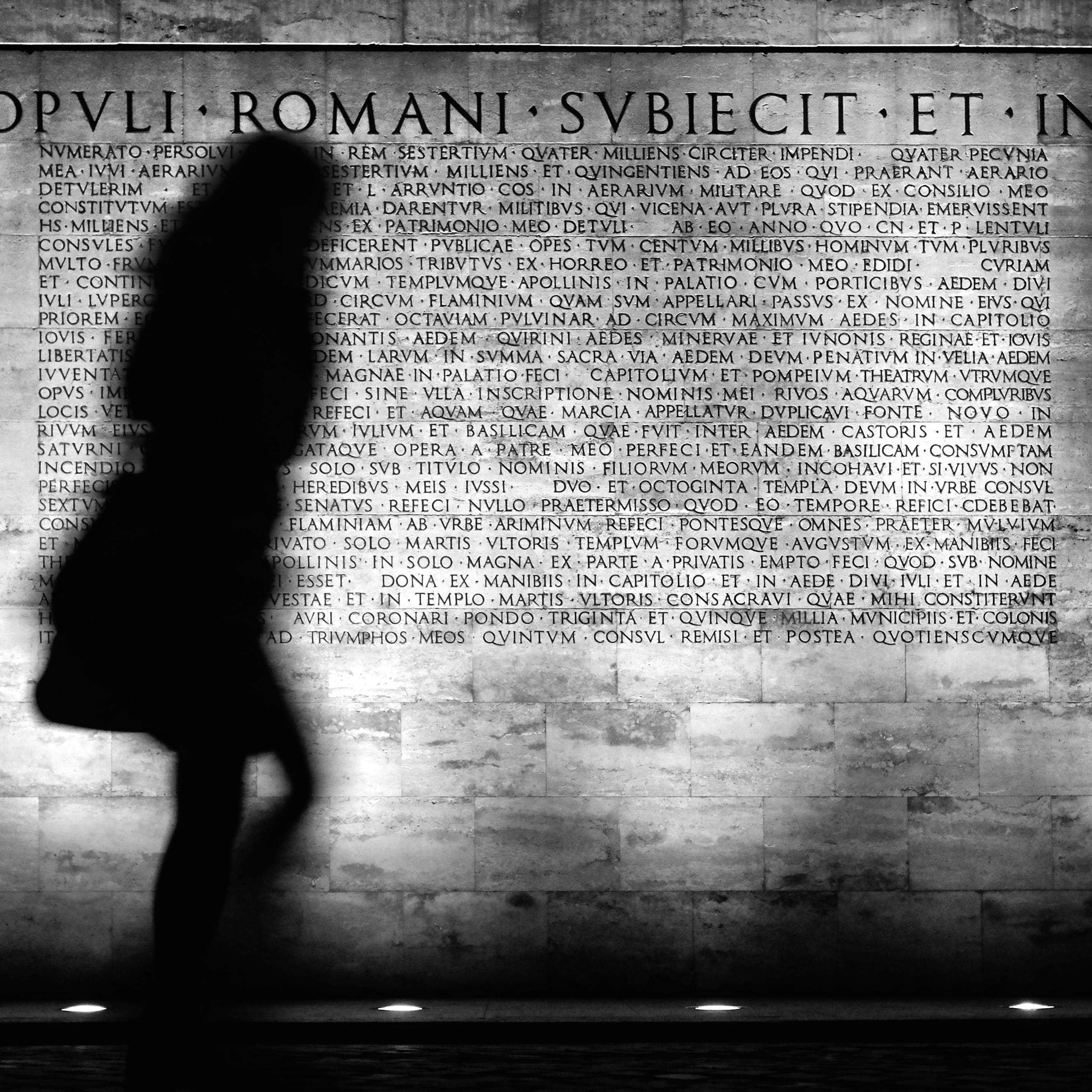 Black and white photo of a large stone wall inscribed with Latin text in Roman capital letters. The inscription is the Res Gestae Divi Augusti (The Deeds of the Divine Augustus), a first-person record of the life and accomplishments of the first Roman emperor, Augustus. A sharp, dark silhouette of a person walking past is cast across the left side of the inscription, partially obscuring the text.