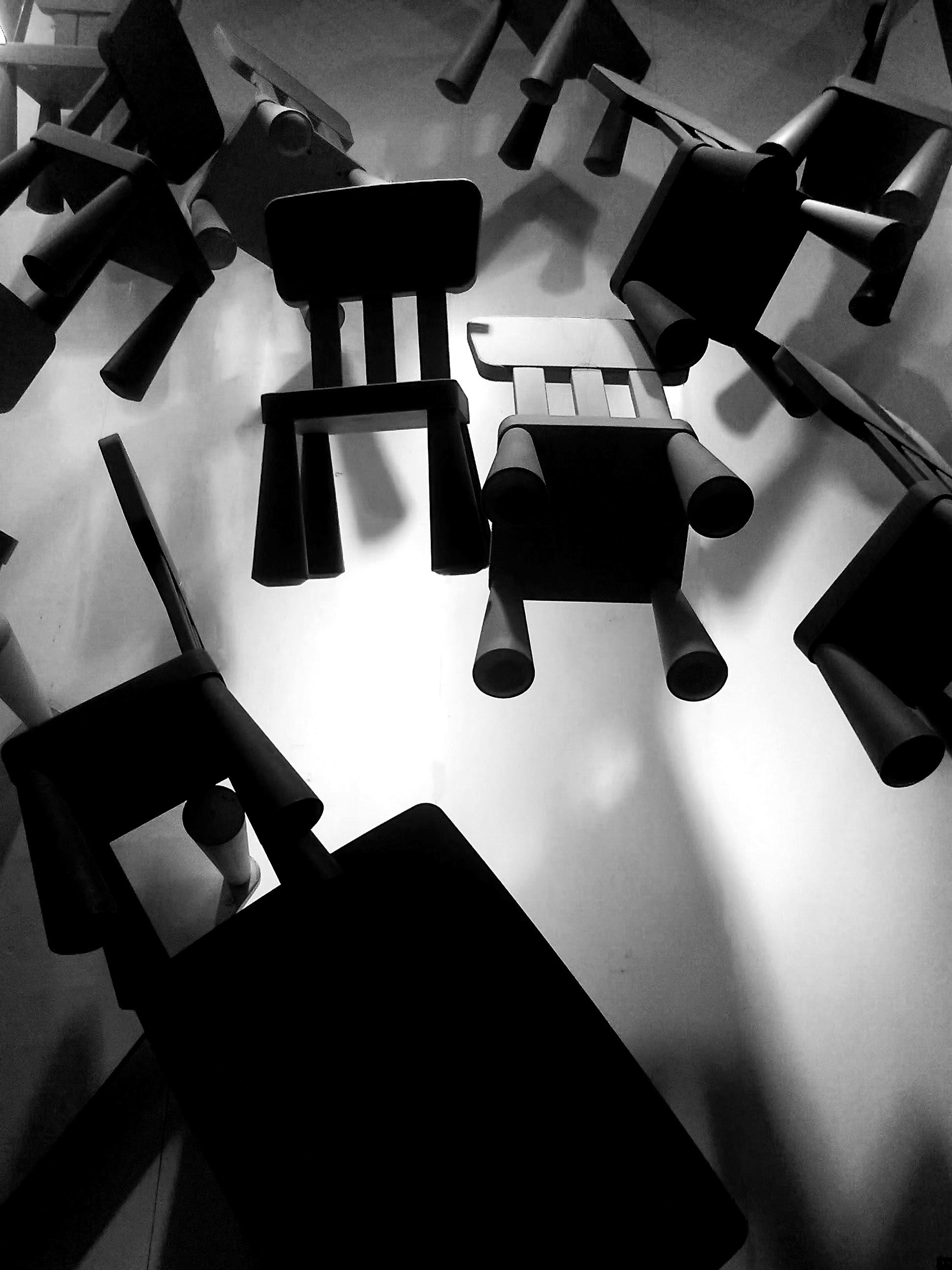 Black and white photo taken from a low angle looking up at numerous plastic chairs suspended from the ceiling. The chairs are arranged in a circular pattern, creating a disorienting, gravity-defying effect. Strong shadows are cast on the white ceiling, emphasizing the geometric shapes of the chair legs and seats.