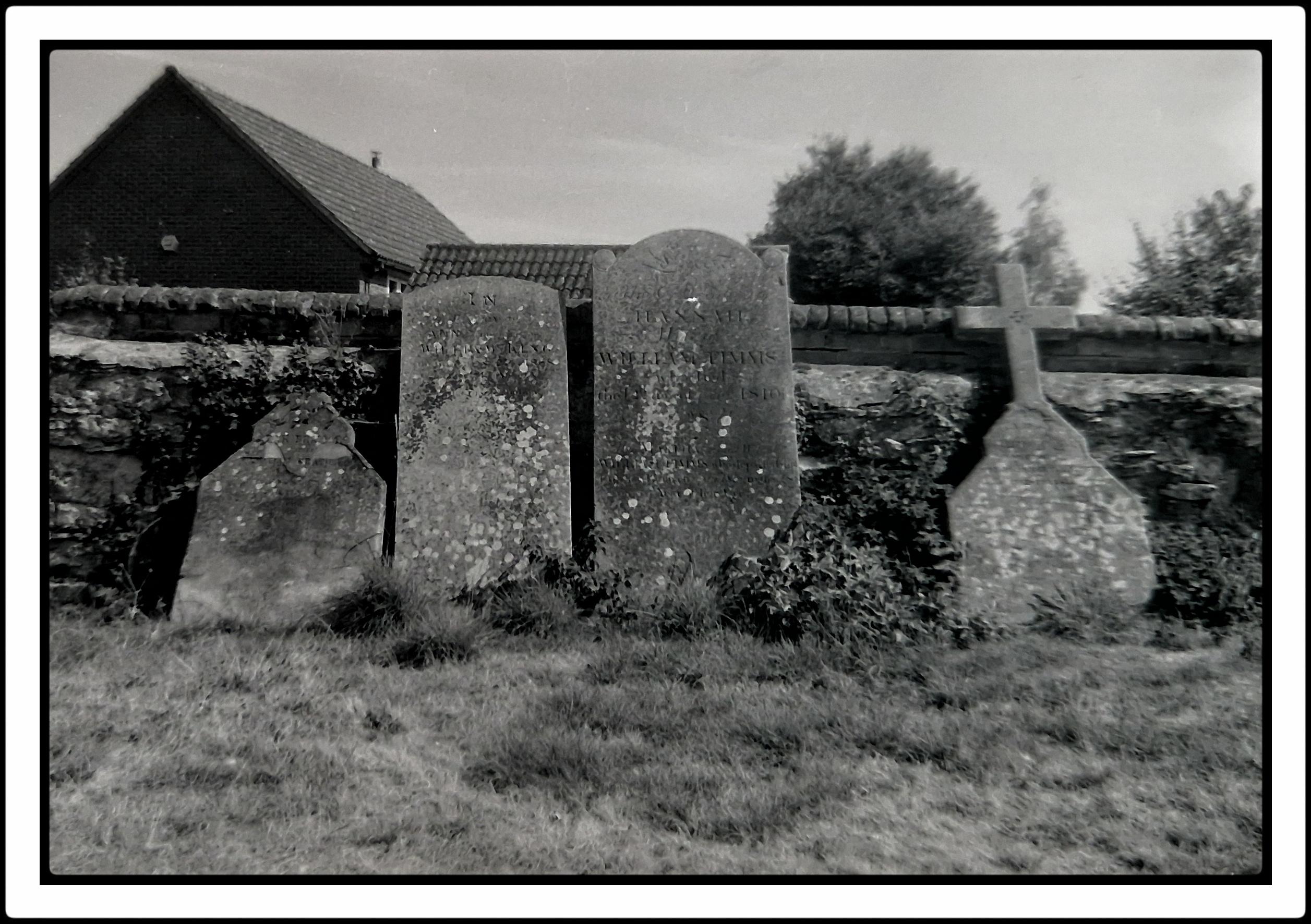 A grainy analogue photo of old gravestones leaning against a wall. Blurred edges.
