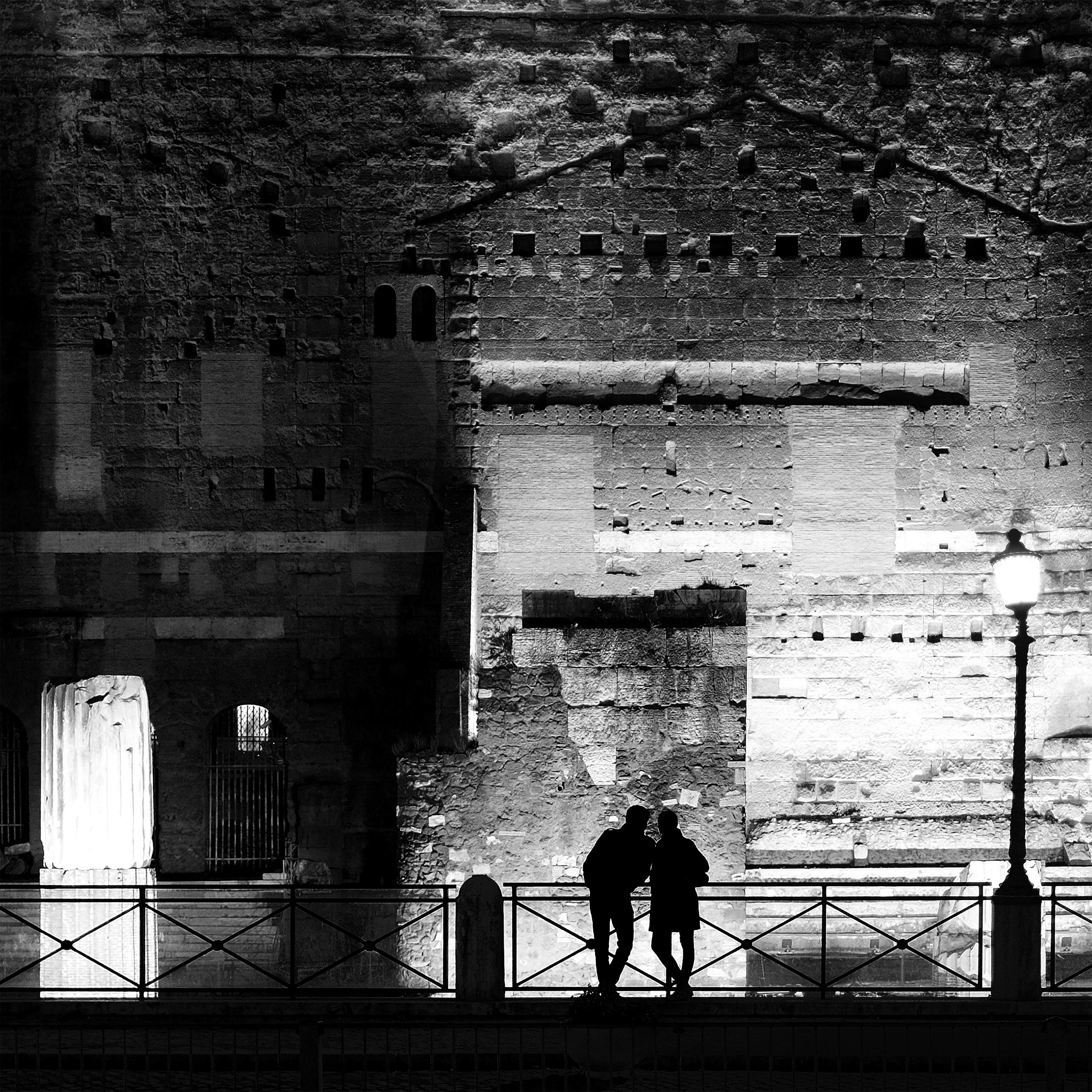 Black and white night photo of a couple in silhouette standing close together against a railing. Behind them is a large, textured ancient stone wall of the Roman Forum, with visible brickwork, arches, and a broken column lit from below. A single lit streetlamp stands to the right. The scene is high contrast, emphasizing the shadows of the couple against the illuminated historic architecture.