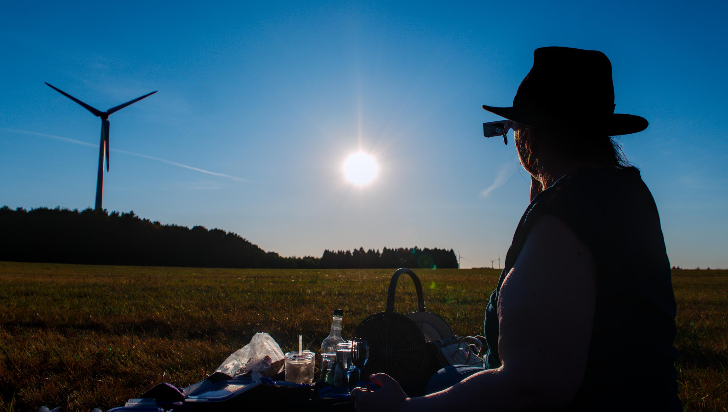 Silhouette von @tvpin beim Picknick auf einer Wiese, während sie durch eine Sonnenfinsternis-Brille schaut. Obwohl die Sonne zu 60% bedeckt ist, sieht man sie im Bild als leuchtenden Kreis. Im Hintergrund sieht man die Silhouette eines Windrads, im Vordergrund unten Umrisse des Proviants.