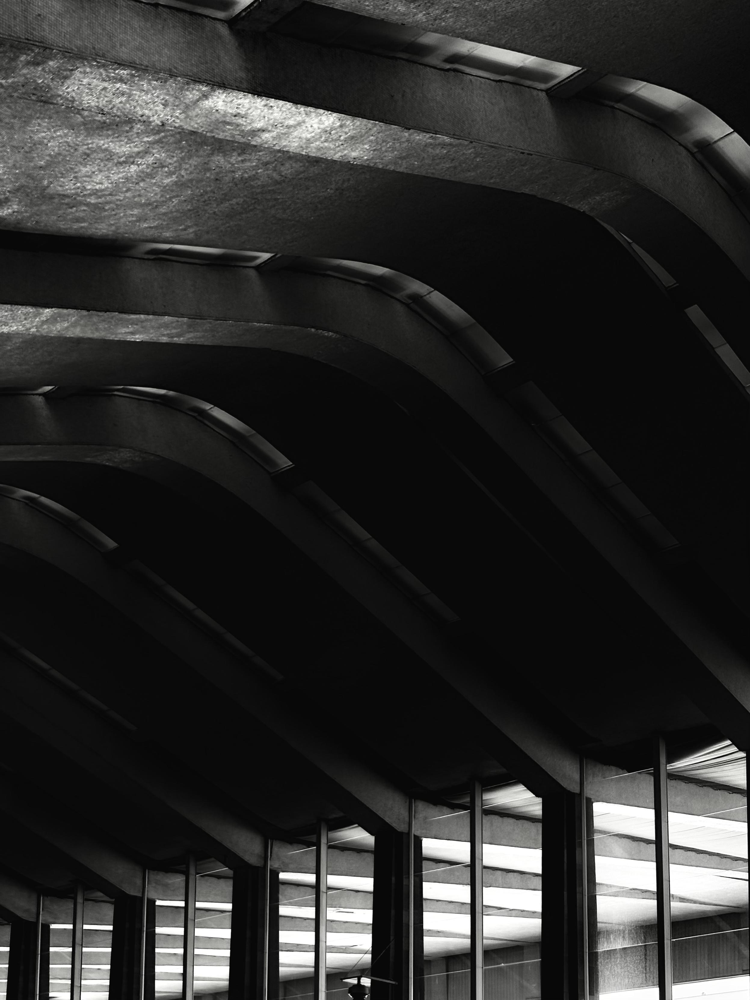 Black and white photo of the curved concrete beams forming the ceiling at Roma Termini train station. The repeating arches create deep shadows and strong diagonal lines, emphasizing the structure's rhythmic, wave-like design. Glass panels below reflect the light, adding depth to the architectural composition.