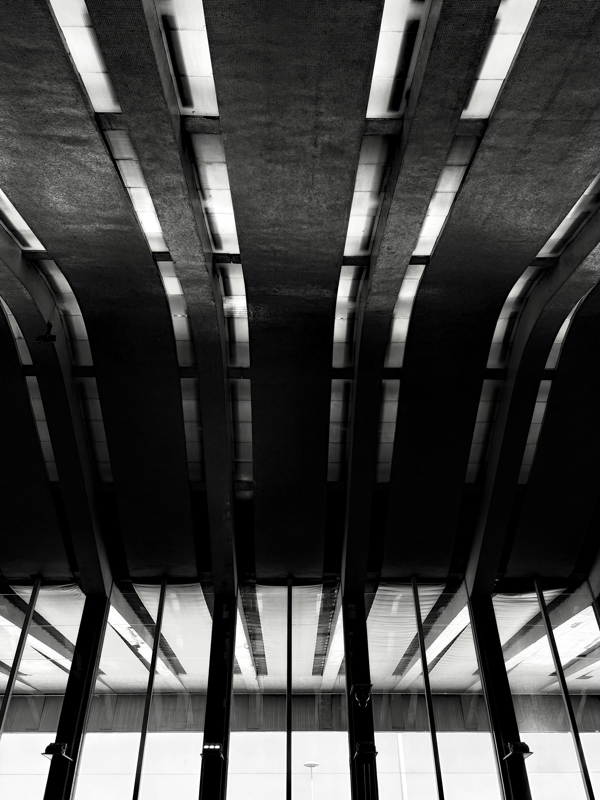 Black and white photo looking up at the ceiling of the main hall at Roma Termini train station. The image shows a series of massive, parallel concrete beams with integrated strip lighting, creating a strong rhythmic pattern of light and shadow. The perspective emphasizes the grand scale and brutalist architectural style of the structure. Large glass panels are visible at the bottom, reflecting the ceiling's geometric lines.