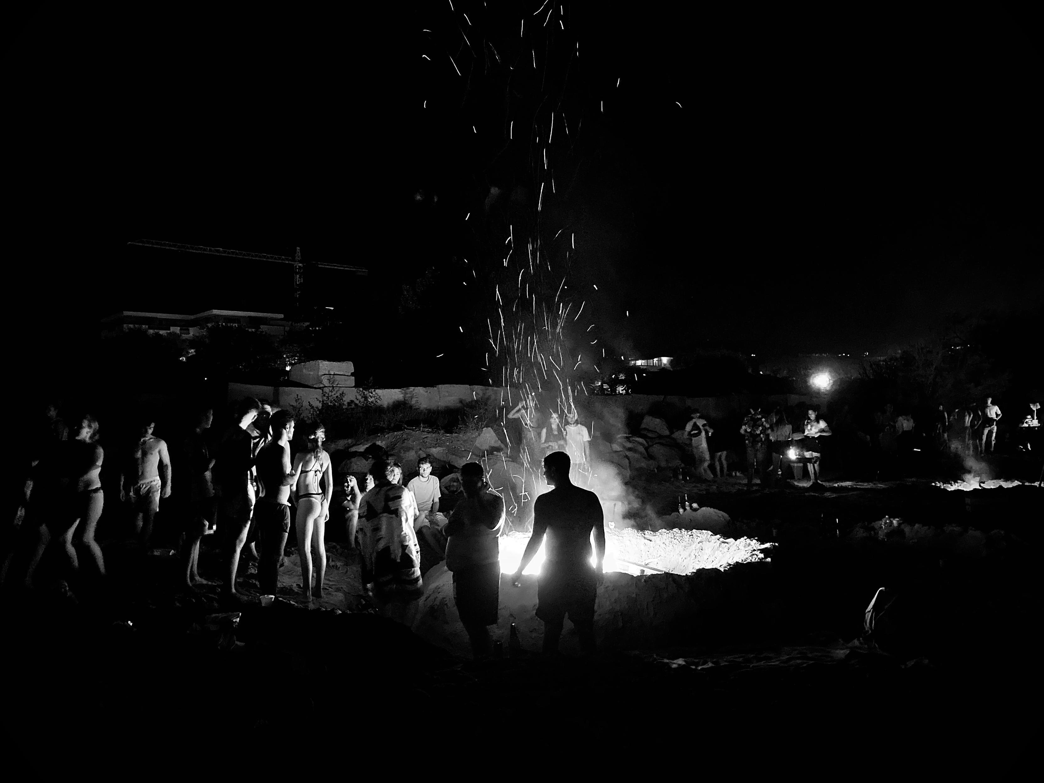 Black and white photo of a group of young people gathered around a large bonfire on a beach at night, with sparks rising into the dark sky.