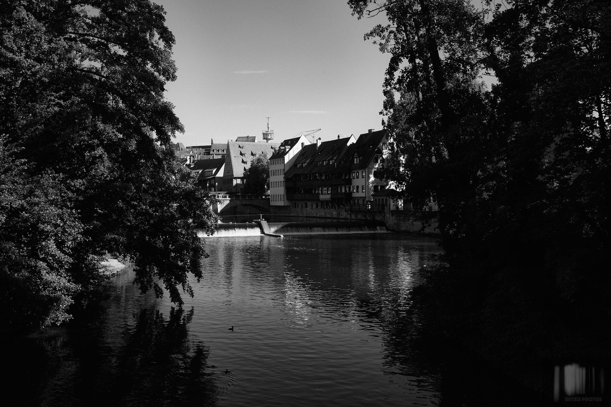 Black and white photograph taken from the Kettensteg footbridge in Nuremberg, showing the old town across the Pegnitz river, framed by dark overhanging tree branches on both sides. A small weir creates white water in the middle distance, with half-timbered buildings visible along the riverbank.