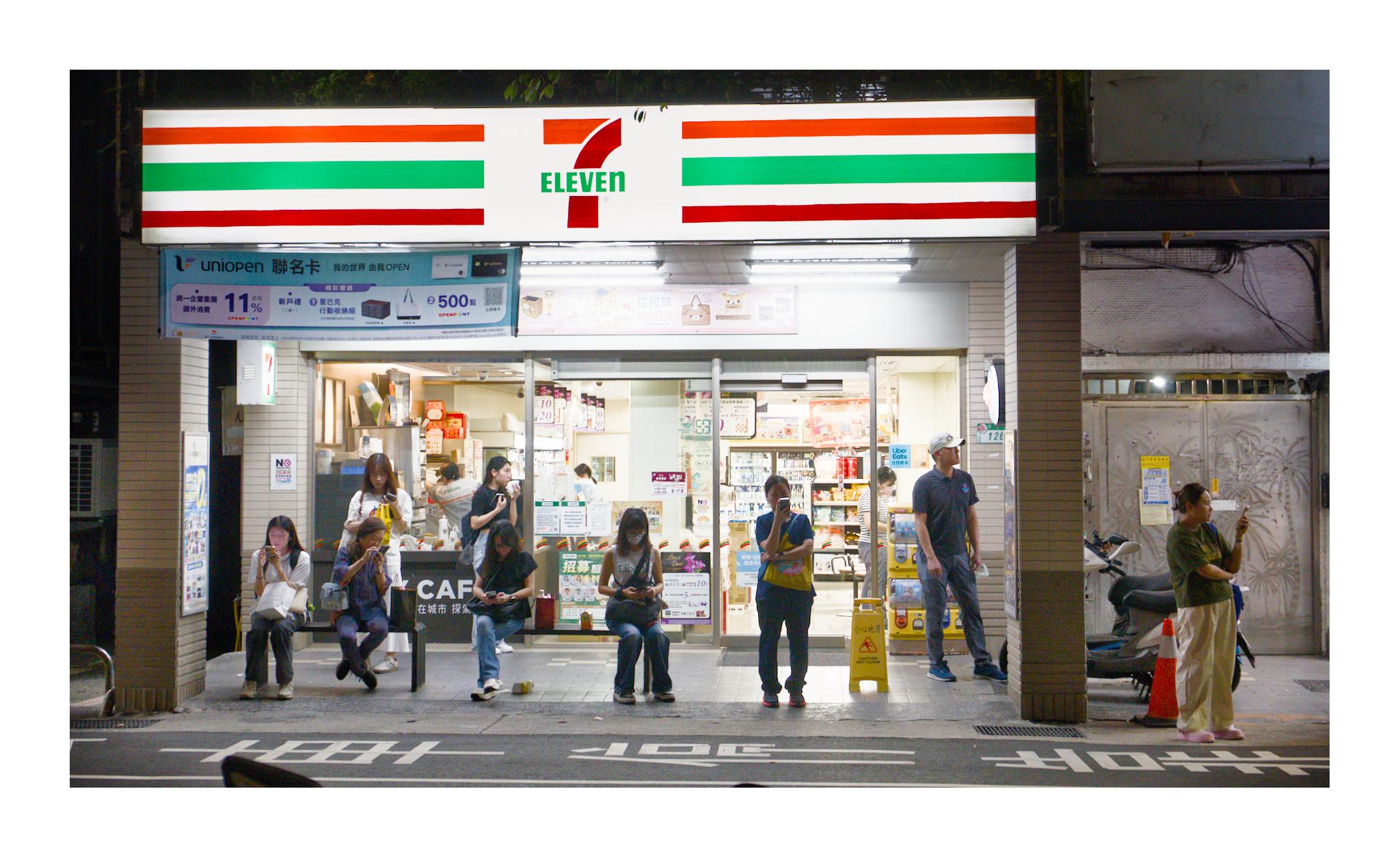 A nighttime street scene in Taipei shows several people waiting outside a brightly illuminated 7-Eleven convenience store. Most are seated or standing separately while looking at their phones. The fluorescent storefront contrasts sharply with the dark street and surrounding buildings, creating a quiet portrait of everyday urban life.