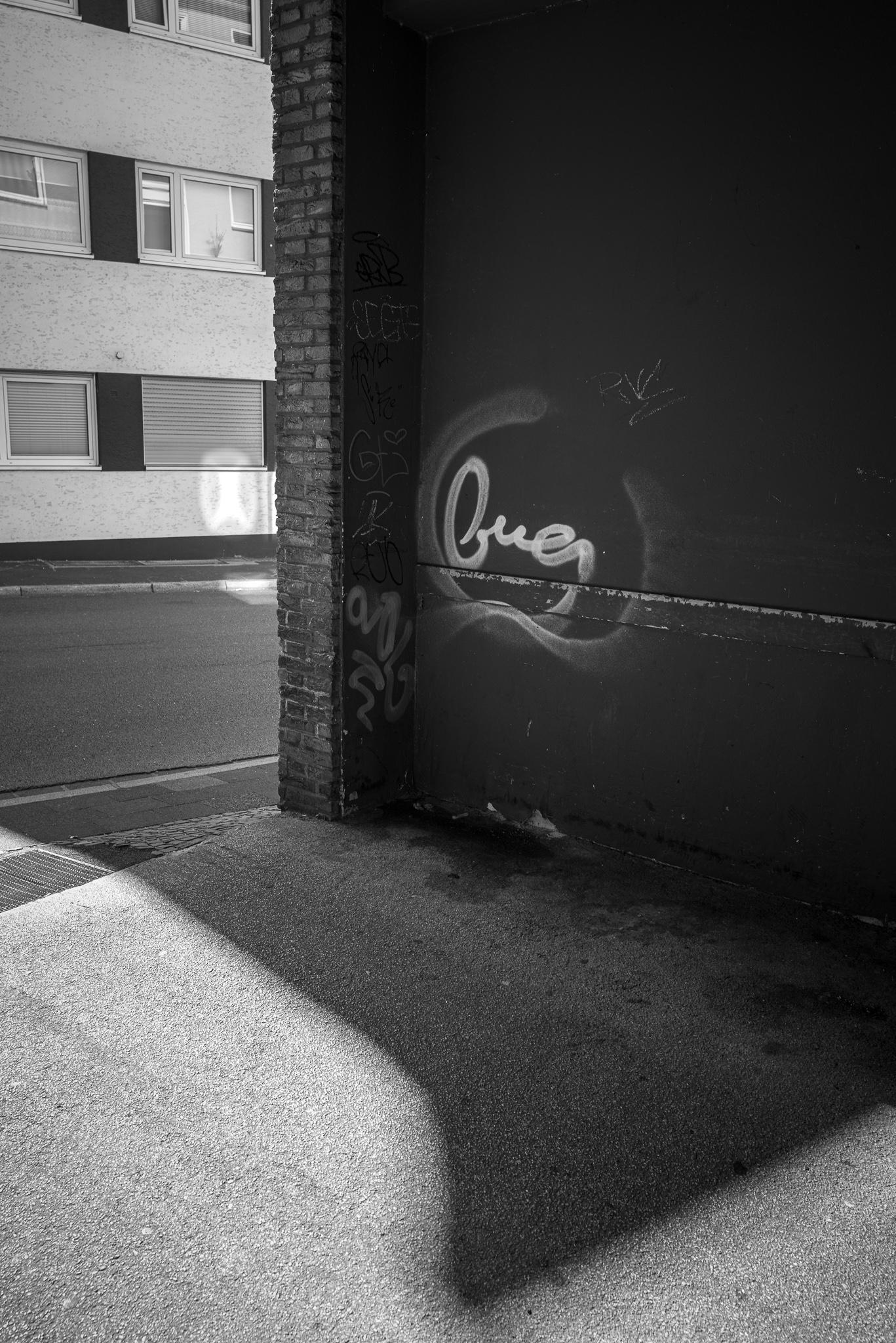 Graffiti saying "bug" in the entryway to a backyard, a shadow falls into the entryway - opposite building is illuminated by patches of reflected light.
