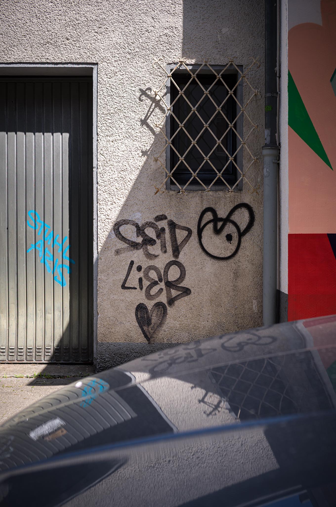 graffiti and shadow and their reflections on the hood of a car parking in front of them, graffiti tag saying be kind in German
