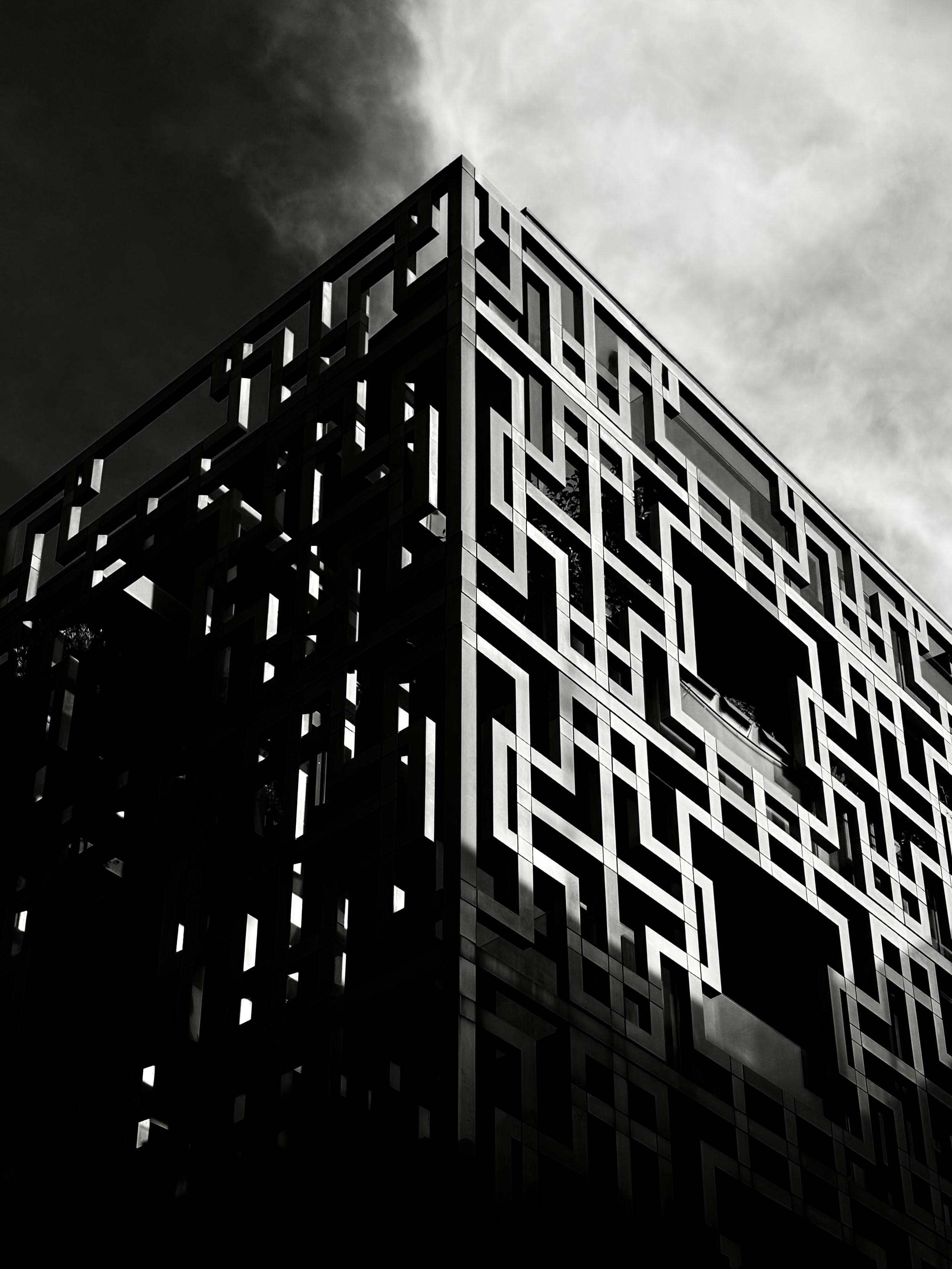 Black and white photo of a modern building with a geometric patterned facade, shot from below with dramatic lighting and shadows.