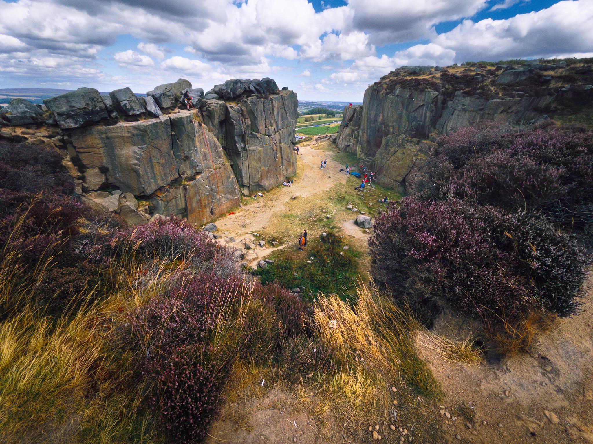 Wide view from the Cow and Calf rocks on Ilkley Moor, looking across a rugged quarry-like gap between tall grey rock faces. Purple heather and golden grasses fill the foreground, while several climbers and walkers are scattered among the rocks and on the sandy ground below. Beyond the cliffs, the landscape opens towards Ilkley beneath a bright blue sky filled with large white and grey clouds.