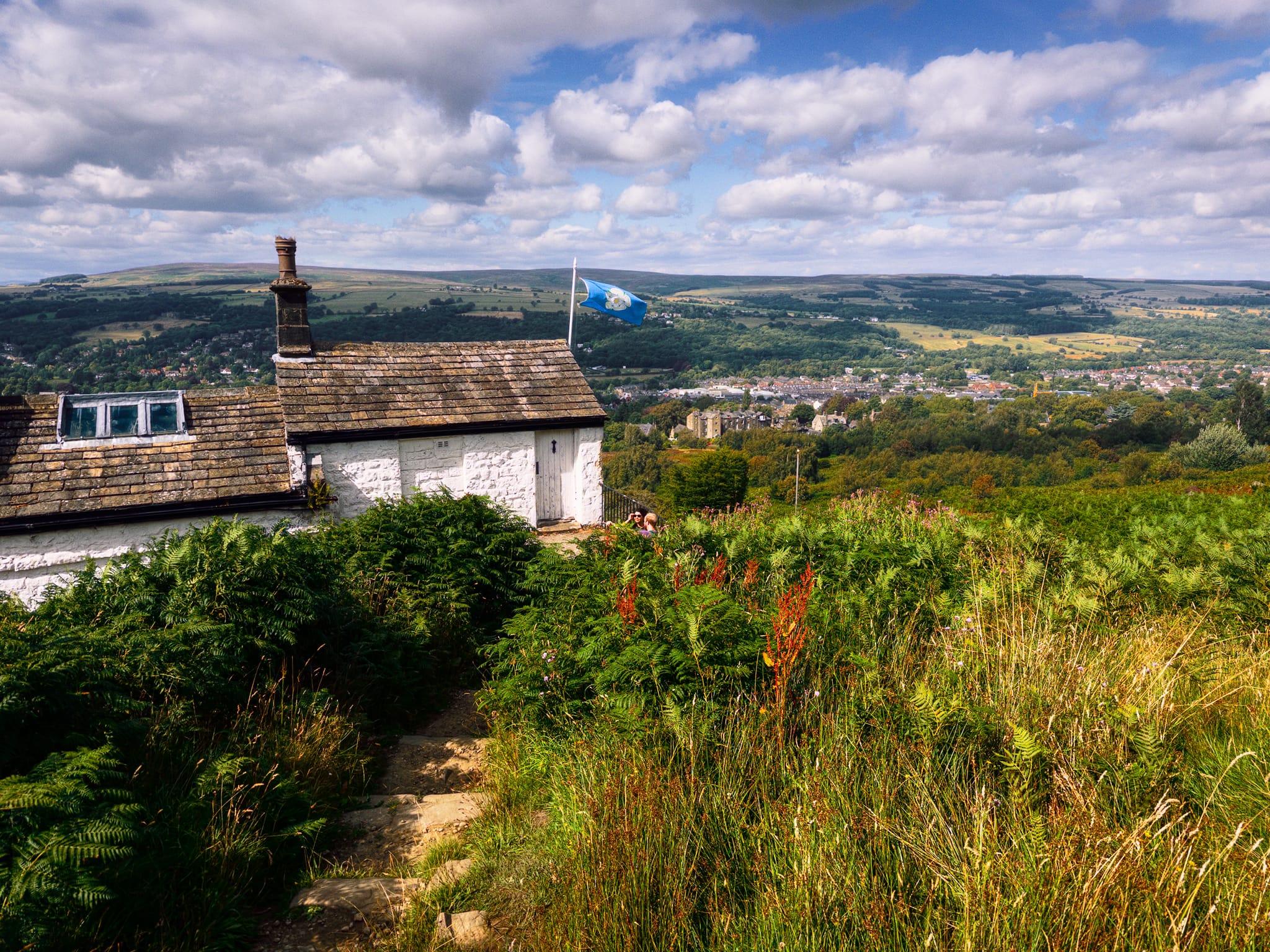 View across Ilkley from White Wells on Ilkley Moor, with the whitewashed stone cottage and its weathered stone roof in the left foreground. A blue flag flies beside the building, while dense green bracken, grasses and wild plants surround a narrow stone path. Beyond the cottage, Ilkley spreads across the valley among trees, with rolling green and brown moorland stretching into the distance beneath a bright blue sky filled with large white and grey clouds.