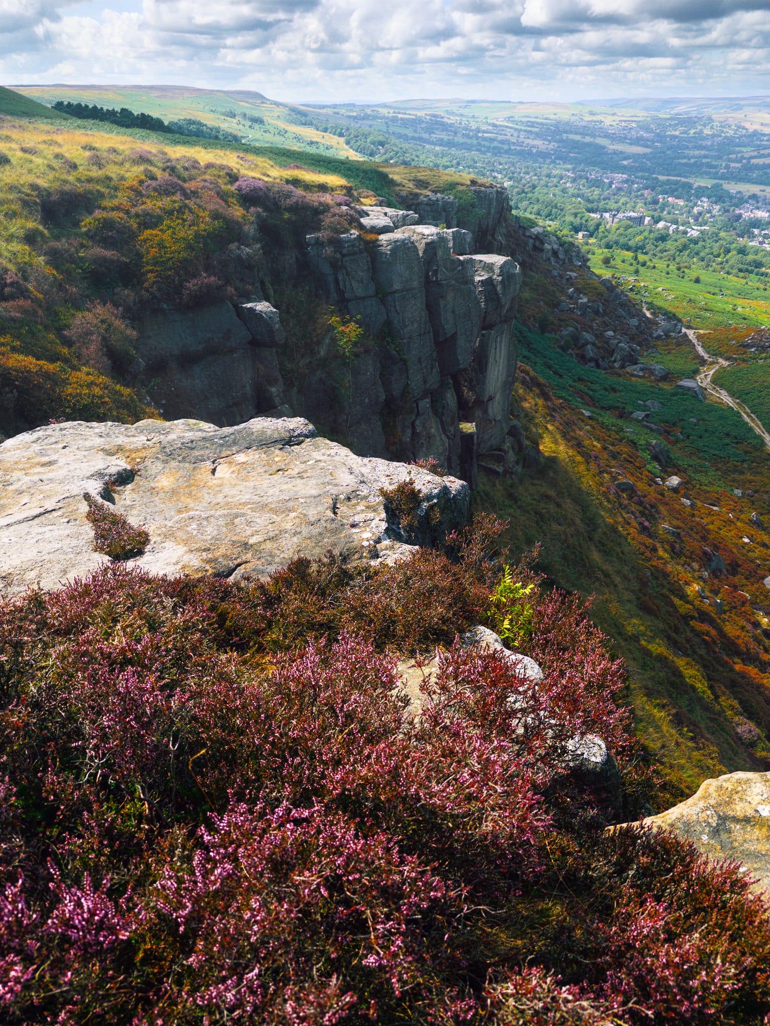 View from Ilkley Crags across a dramatic line of sheer grey rock faces descending into the valley, surrounded by dense purple heather, grasses and patches of green bracken. Pale rocky ledges fill the foreground, while a narrow footpath winds along the lower hillside to the right. Beyond the crags, Ilkley and the wooded Wharfe Valley stretch into the distance, framed by rolling moorland beneath a bright sky scattered with large white and grey clouds.