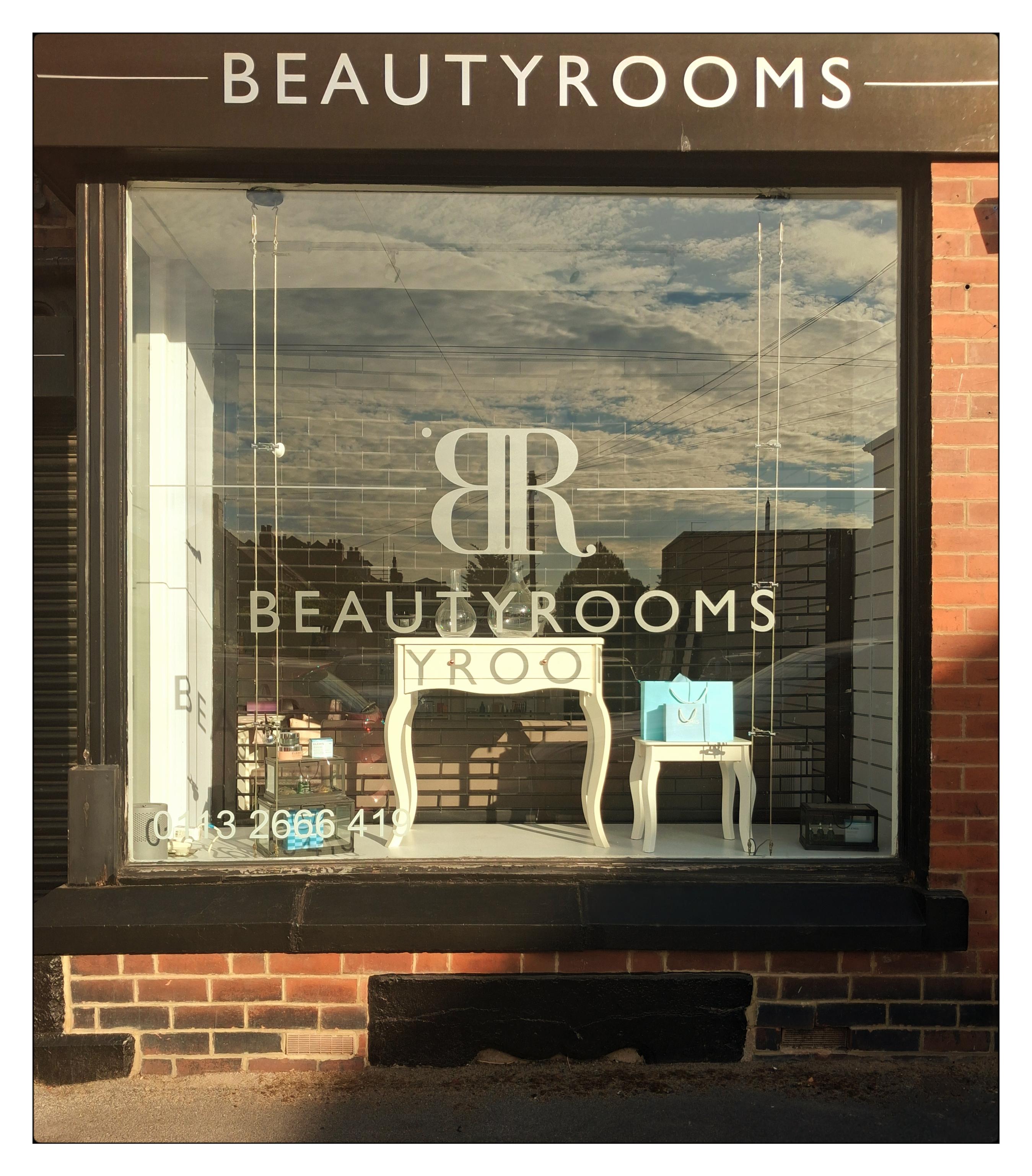 A large square window set in a red brick wall. A shop front. The words 'Beauty Rooms' are on the glass and blue sky and clouds are clearly reflected. The letters YROO are projected in shadow on to a table inside.