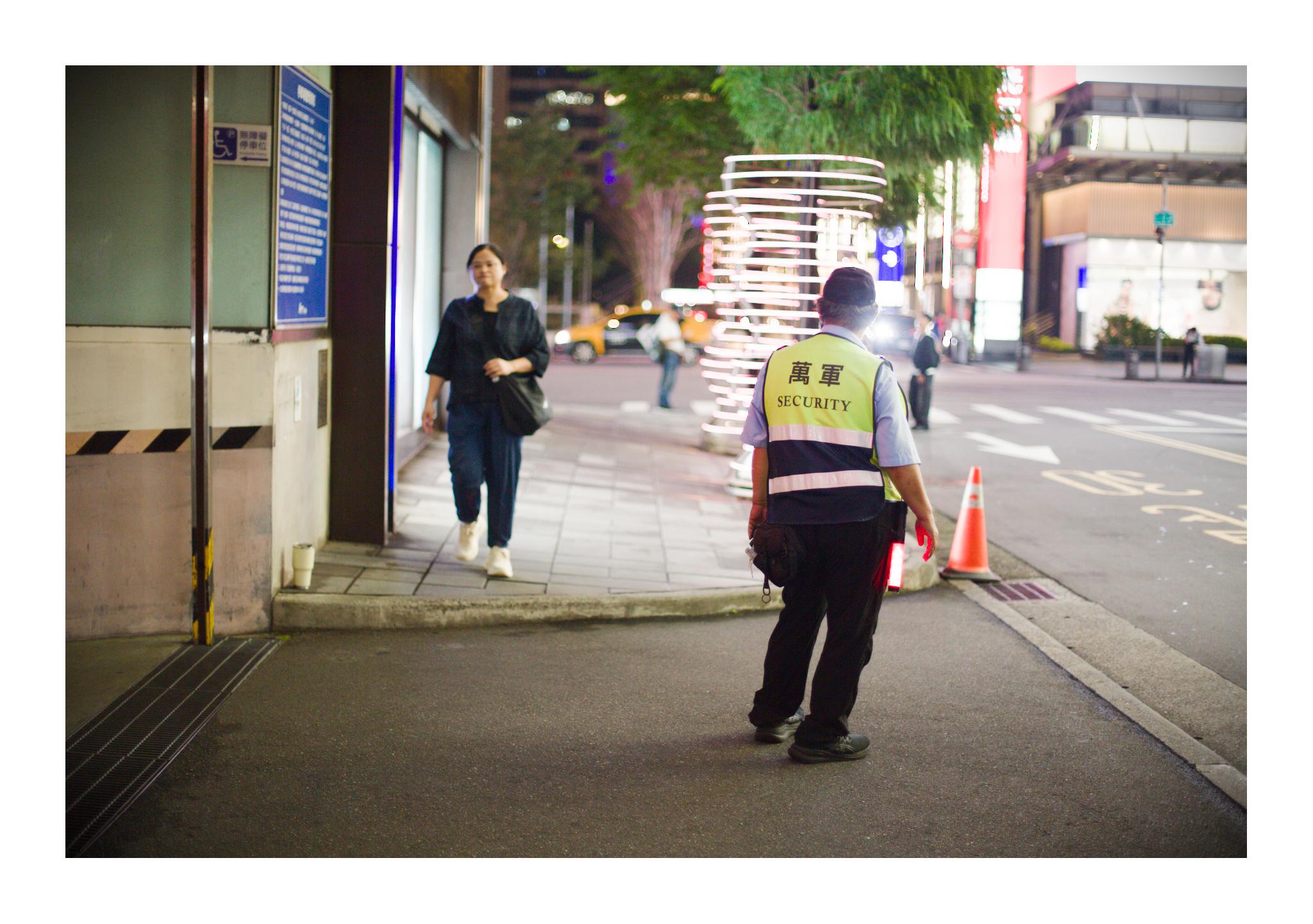 A security guard stands on a brightly lit sidewalk at night in Taipei’s Xinyi District, facing the street. His reflective vest reads “SECURITY” beneath Chinese characters. A woman approaches from the opposite direction, while glowing storefronts, traffic and a large illuminated sculptural structure create a soft, colorful background.