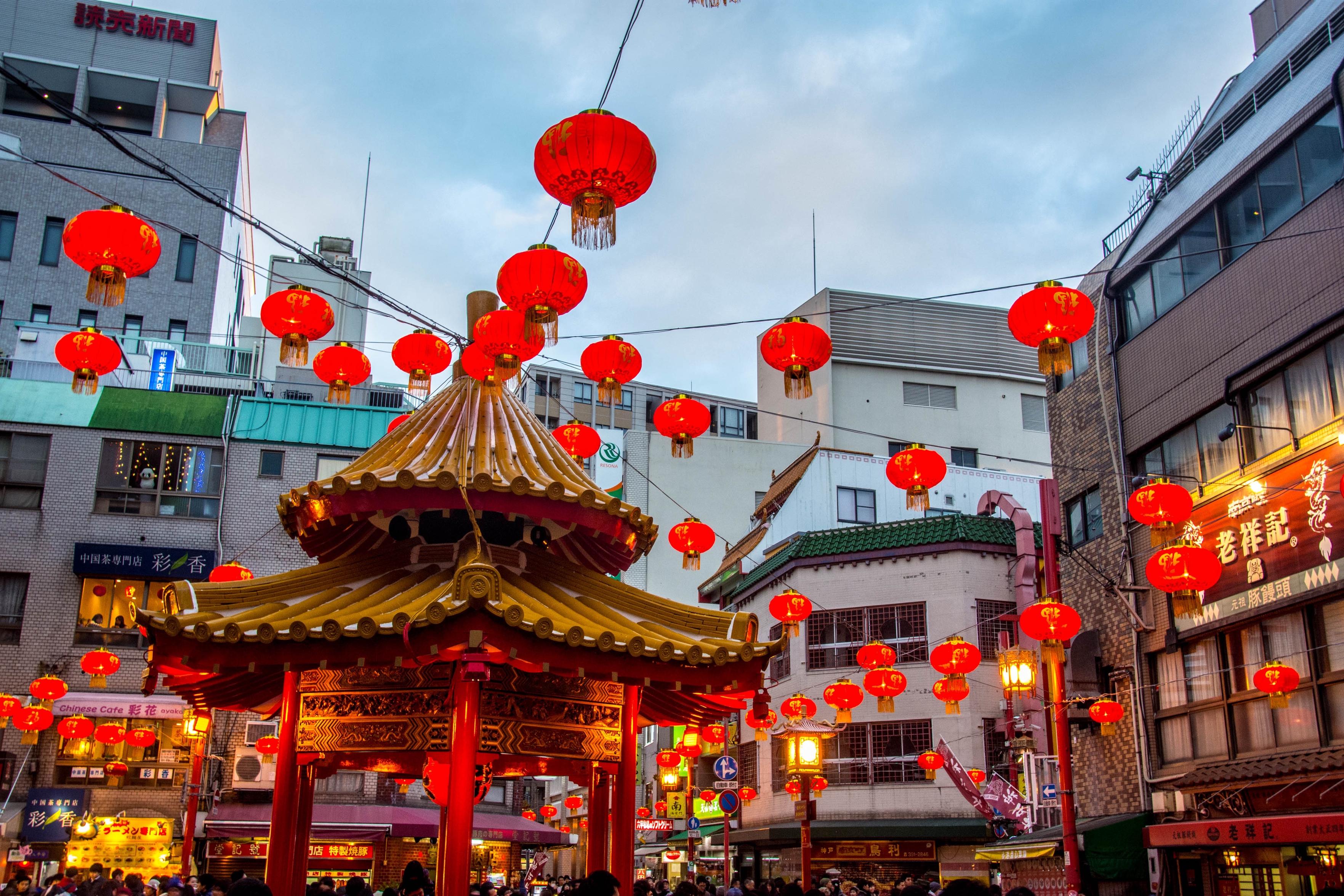 A bustling street adorned with numerous red lanterns, featuring a decorative pavilion in the foreground. Surrounding buildings display a mix of traditional and modern architecture, creating a vibrant atmosphere. The sky is overcast, adding to the scene's charm.