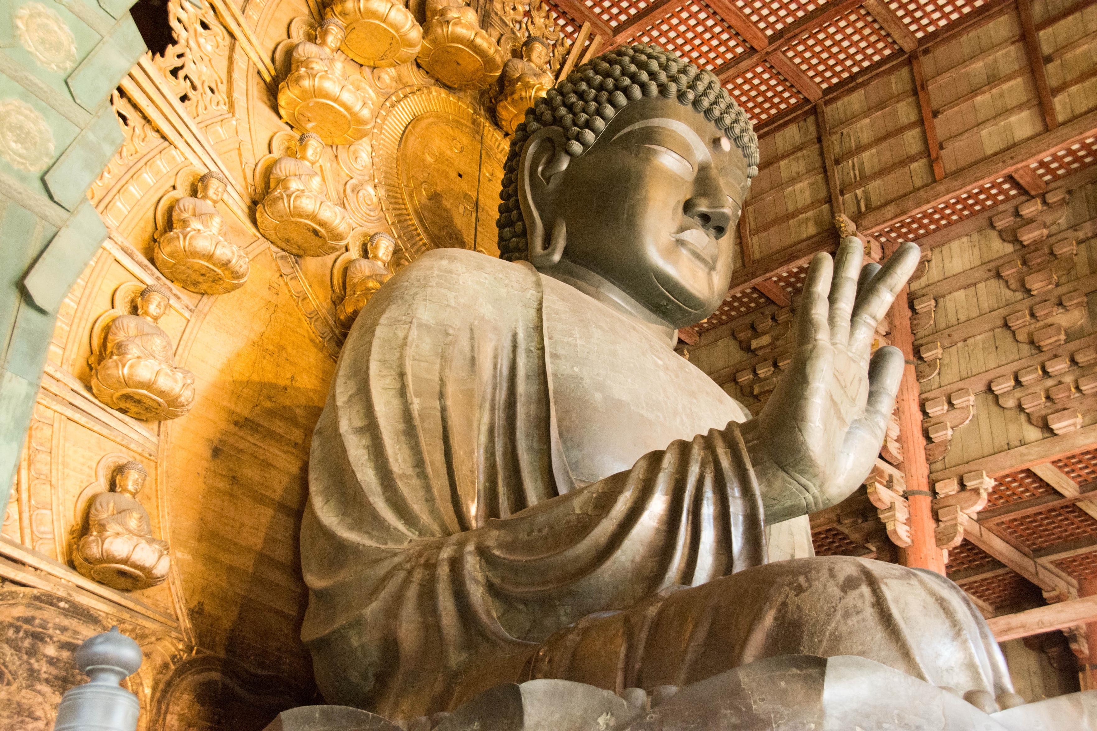 A large statue of Buddha sits peacefully, adorned with intricate details. The background features ornate decorations with smaller buddha figures set against a golden wall, under a wooden ceiling. The statue's hand is raised in a gesture of peace.