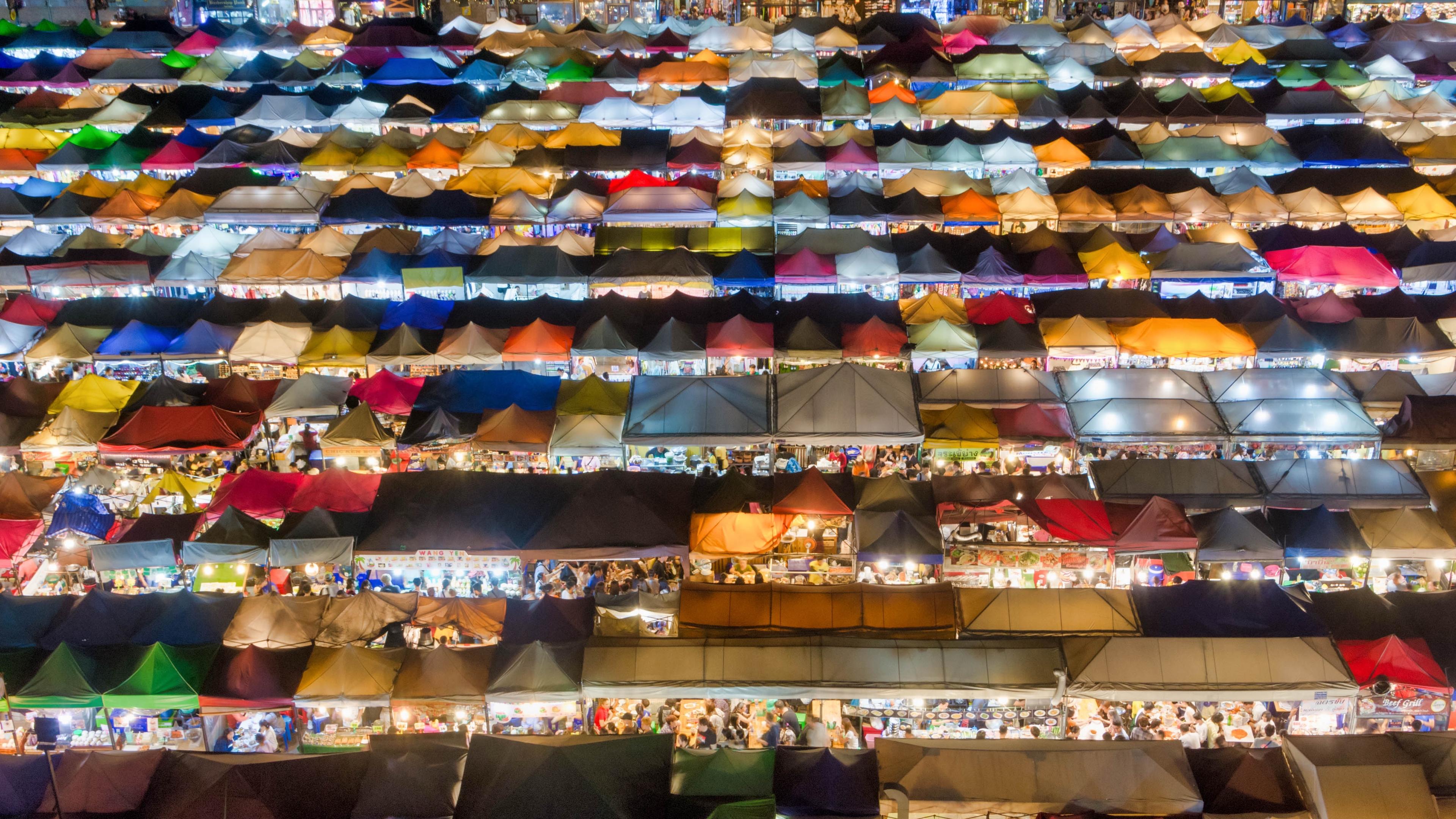 A vibrant aerial view of a bustling night market, showcasing numerous colorful tents filled with vendors and customers. The scene highlights an array of lights and activities across the densely packed stalls.