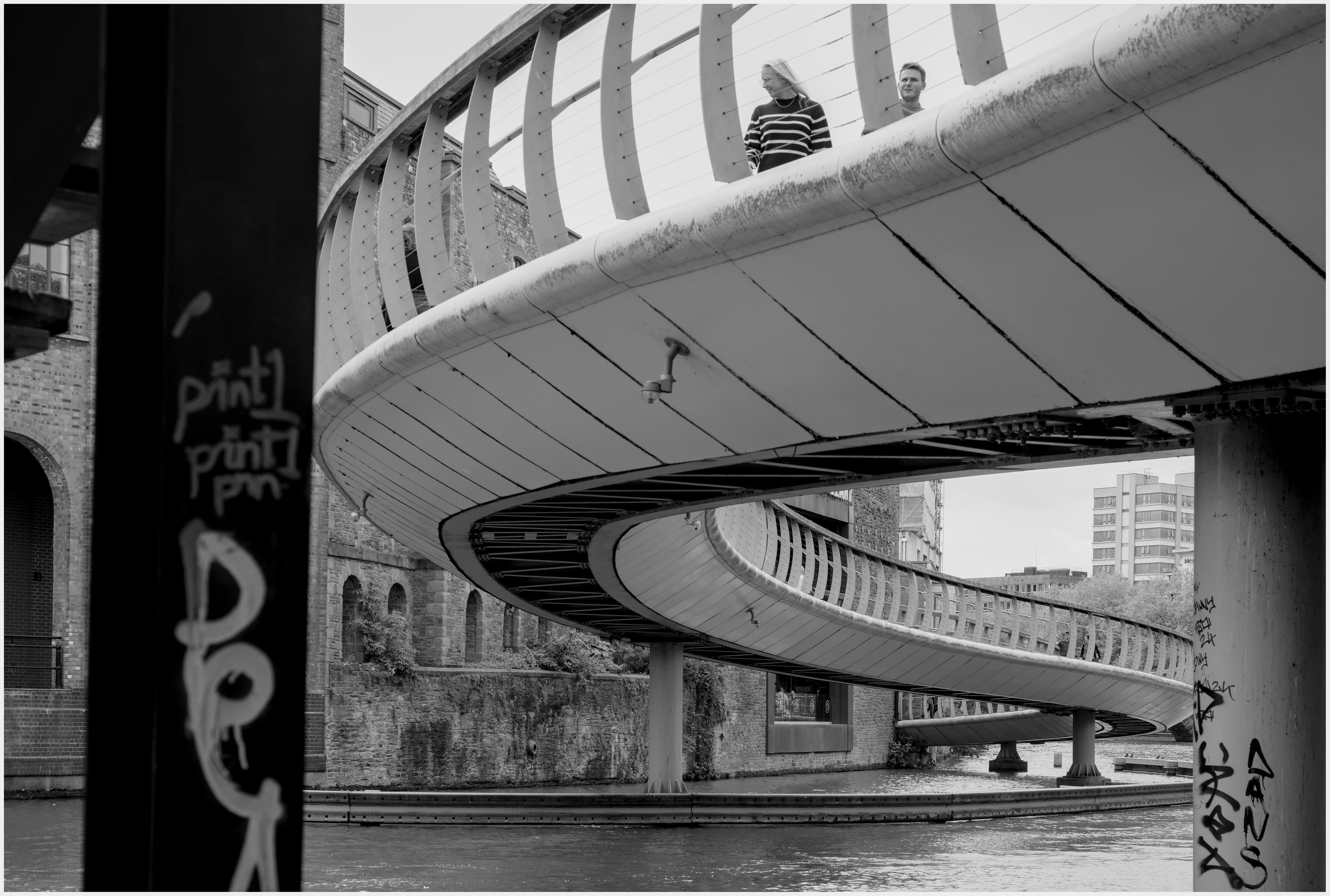 A black and white low angled photgraph of the 'S' Curve footbridge in central Bristol, UK with two people walking on curve nearest the camera. The female is turning and looking away to her right and the male is just turning his head to the right. There is some grafitti on pillars near the flowing river water in the lower half of the image. The lighting is quite flat and over cast though bright so the contrast was difficult to acheive. Why: I liked to strong curves of the bridge and the presence two people for me give the picture context.