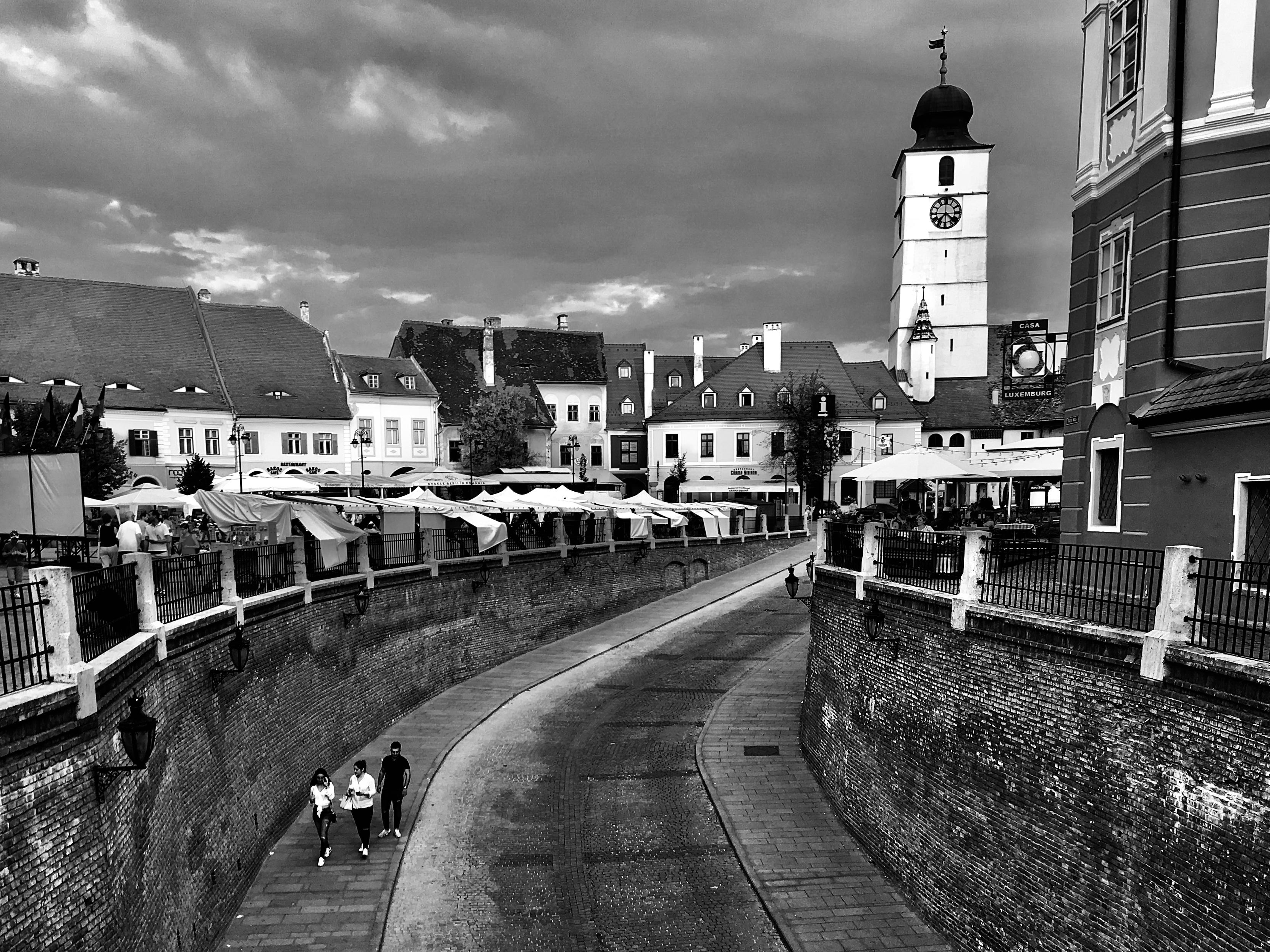 Black and white photo of a sloping cobblestone street in Sibiu, Romania, framed by high brick walls. Three people walk downhill in the foreground. In the distance, the historic square features rows of white market umbrellas and traditional buildings. The tall, white Turnul Sfatului (Council Tower) with its clock and dark dome dominates the skyline against heavy clouds