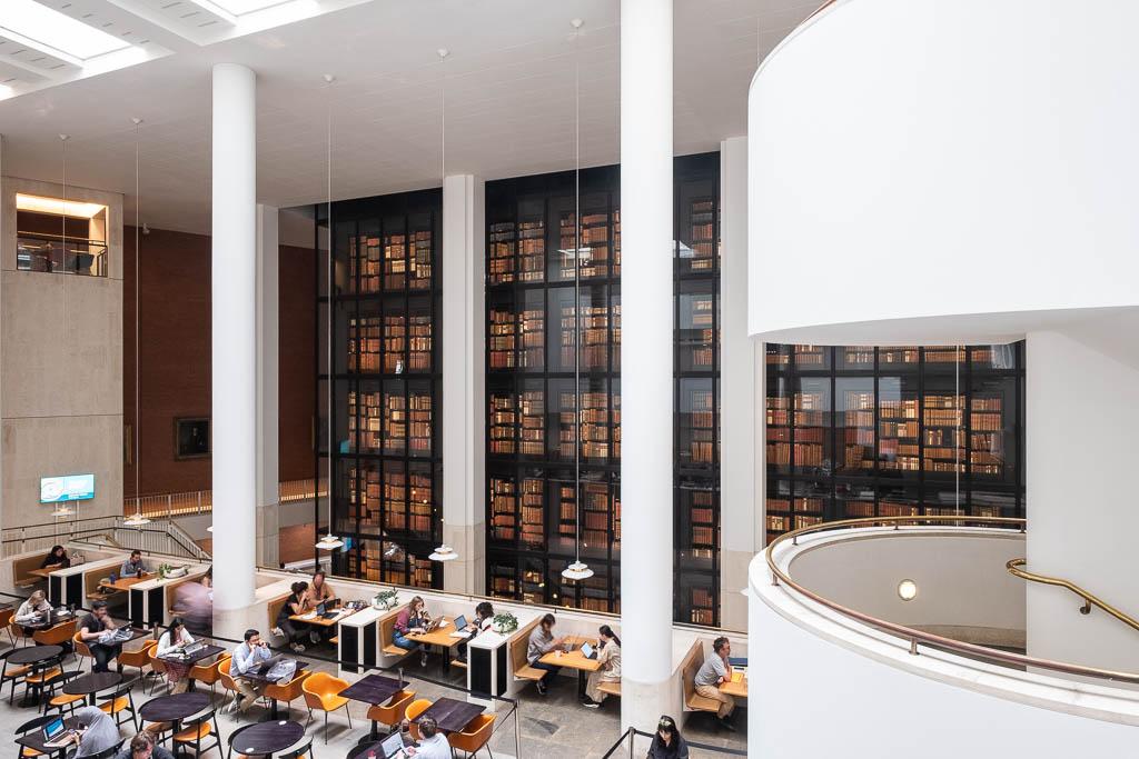 Interior view of a modern library. A white staircase with brass railings occupies most of the forefront, to the right. To the left is a reading room with small, cafe-like tables and patrons working on computers. In the background is a large black glass box filled with rare books that glow in a warm light.
