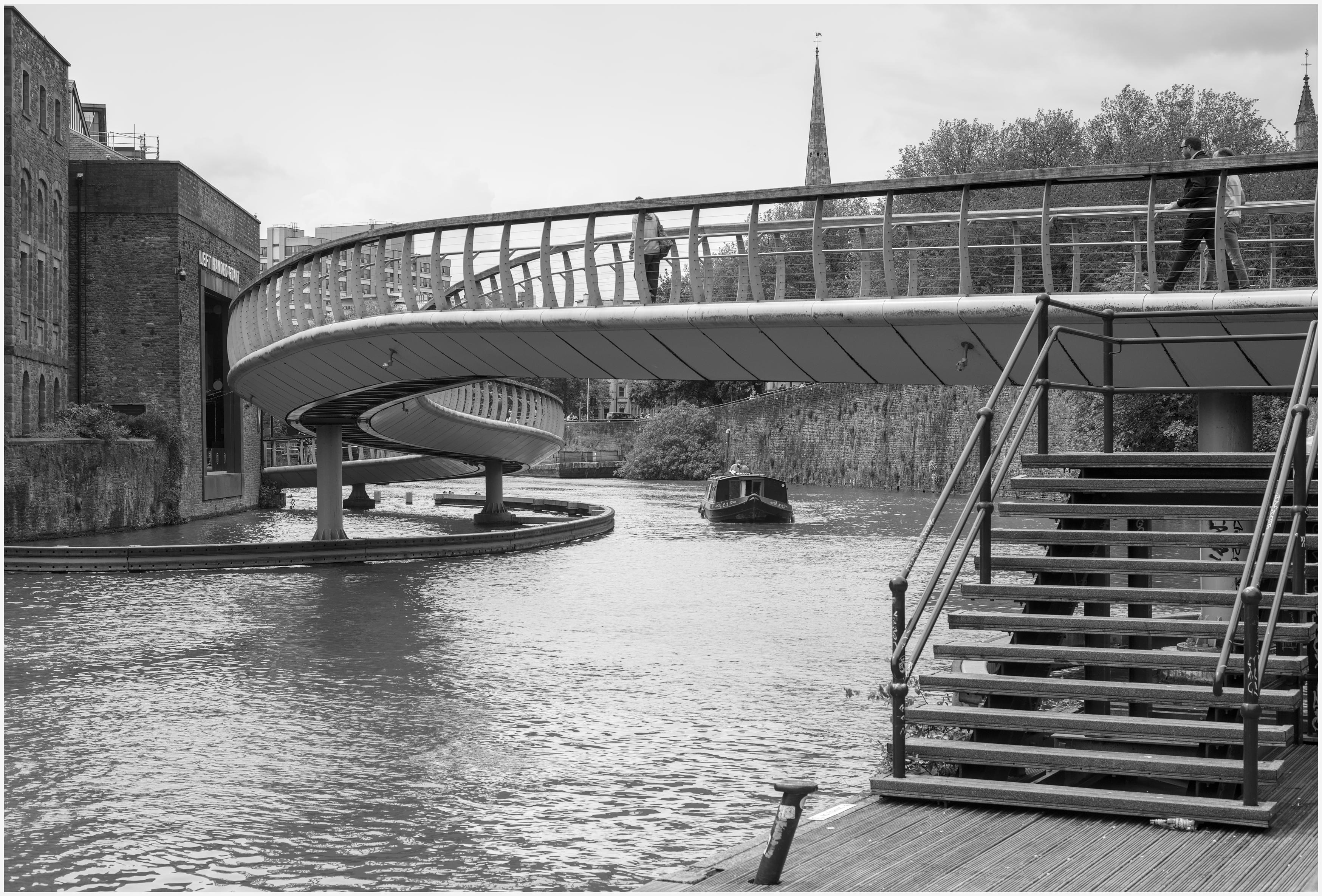 A black and white low angled photgraph of the 'S' Curve footbridge in central Bristol, UK. Photographed from a wooden decked landing stage, accessed via a dozen steps, at the bottom right of the image. People are walking across curved bridge, though only the head of of one man can be clearly seeen. A river boat, near the middle of the frame, is chugging towards the landing stage. 
A fairly bland overcast sky with some cloud for relief and the lighting is quite flat and over cast, though bright, so the contrast was difficult to acheive.
This mage is a mix of old Bristol with waterfront square industrial buildings, now converted to apartments (flats) and the more modern 'S' Bridge's curves that mimic the bends of the river.