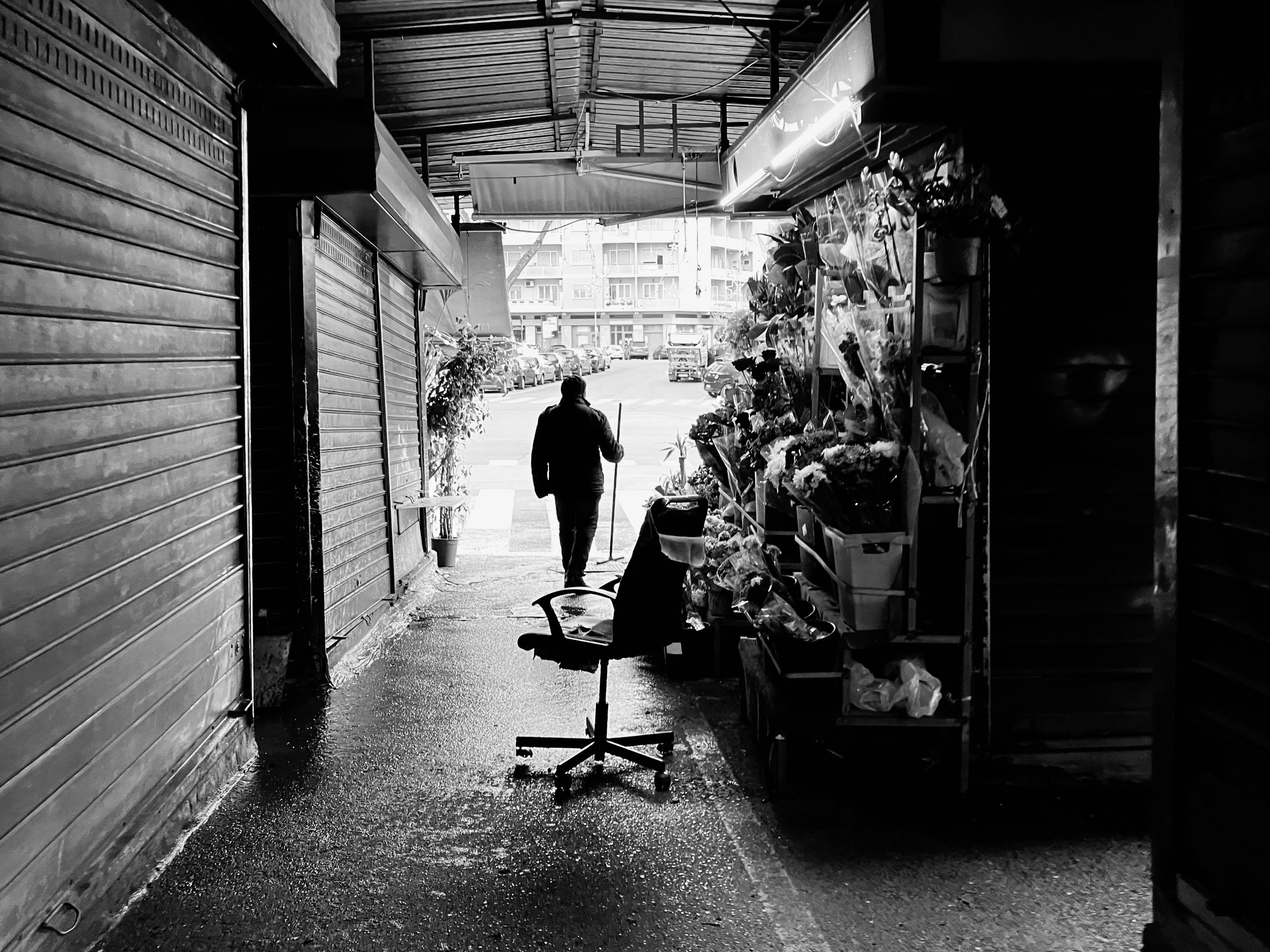 Black-and-white photo of a covered market alley. An empty office chair sits in the foreground next to a flower stand. A lone figure stands holding a broom, facing a sunlit street with parked cars