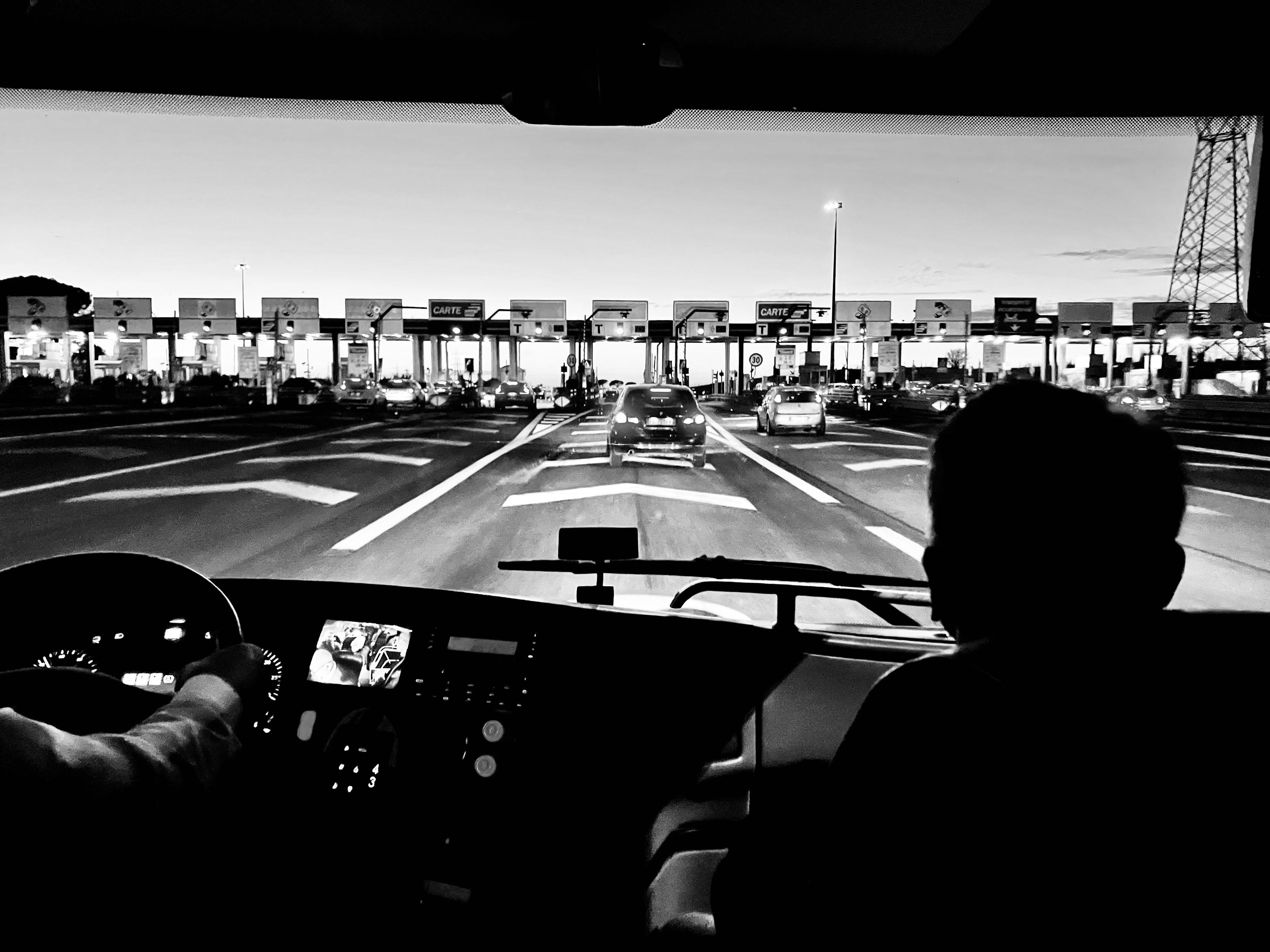 Black-and-white view from inside a bus approaching a toll booth in Rome, with the driver’s silhouette and dashboard visible