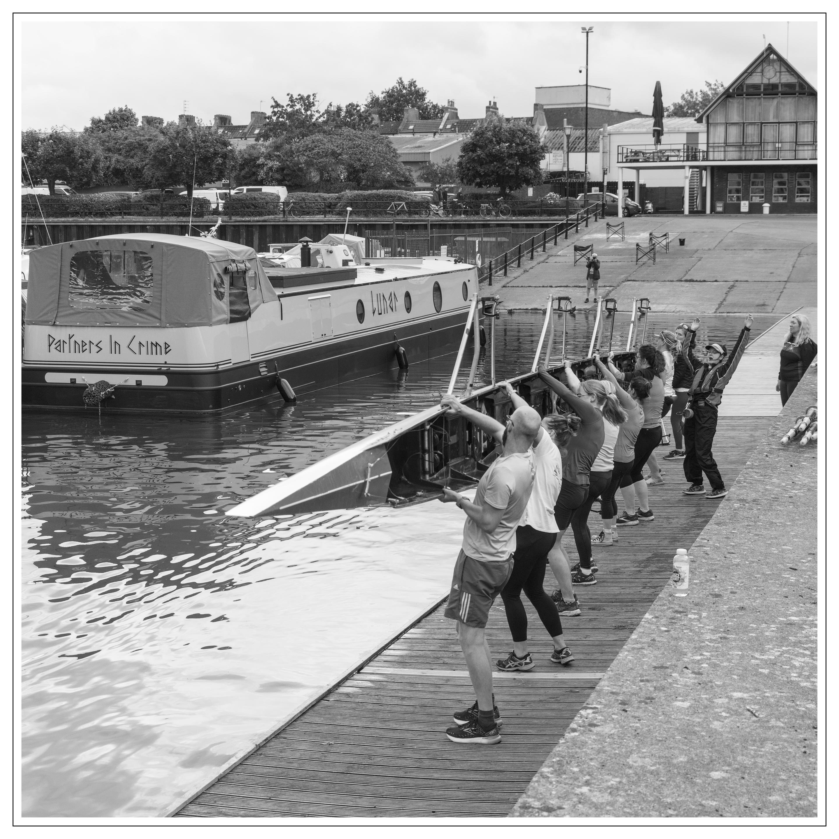 A black and white evening photgraph taken at Bristol Harbour, UK.
The image is an action picture of a Coxed Eight Shell being lifted up by its male and female crew, whilst the the Coxswain encourages them, by flinging her arms up in the air and, disconcertingly, closing her eyes.