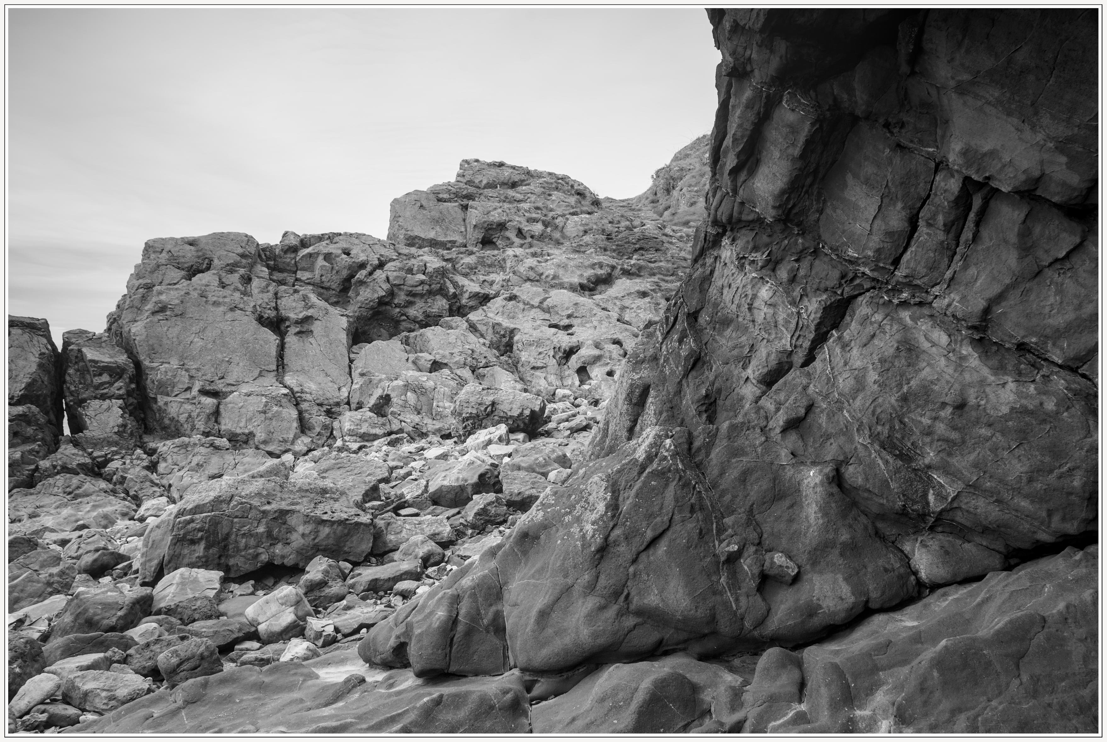 A black and white photgraph taken on the shoreline at Birkett Road, Weston-super-Mare, UK.
The image is of rock formations at the waters edge of the Bristol Channel, Weston Bay.
Rocks are strewn across the shore, from falls caused by erosion.
At the right of the image, from bottom to top, is a solid formation of rock in the sha